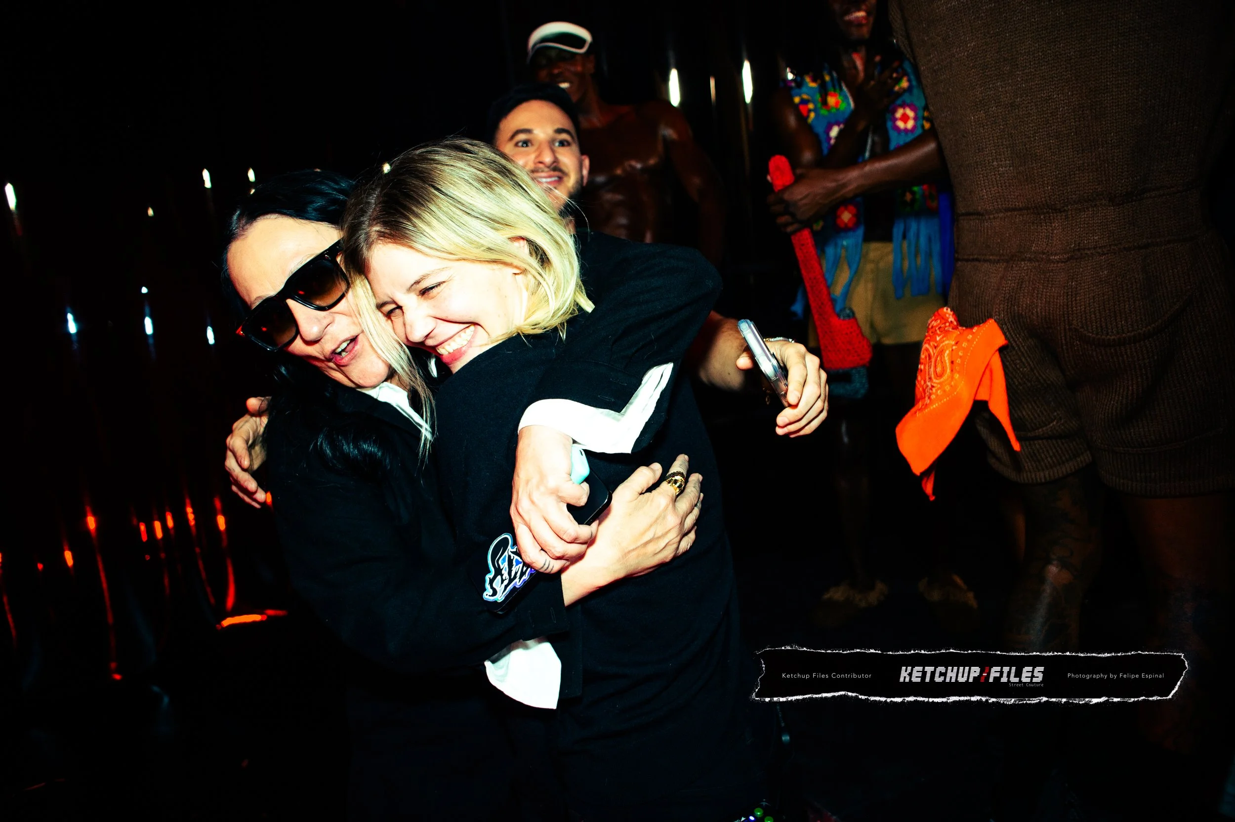 Two women hugging and smiling at a lively party or event, surrounded by other people in vibrant clothing.