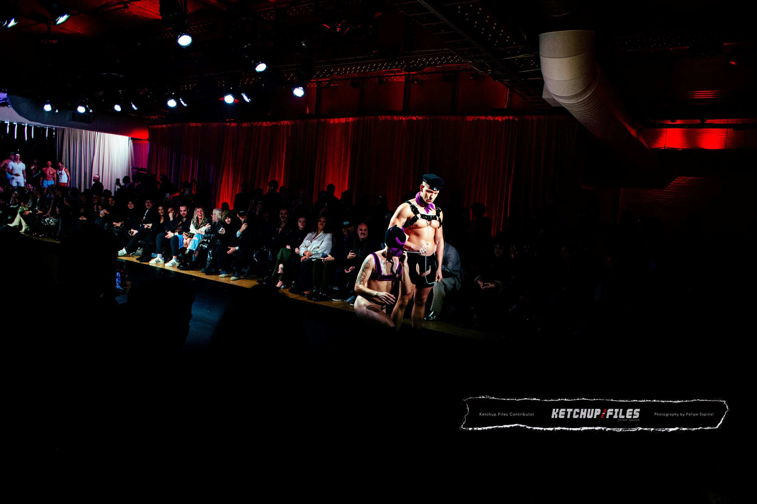 A fashion show runway with two models in provocative outfits, with an audience seated on either side, and stage lighting illuminating the scene.