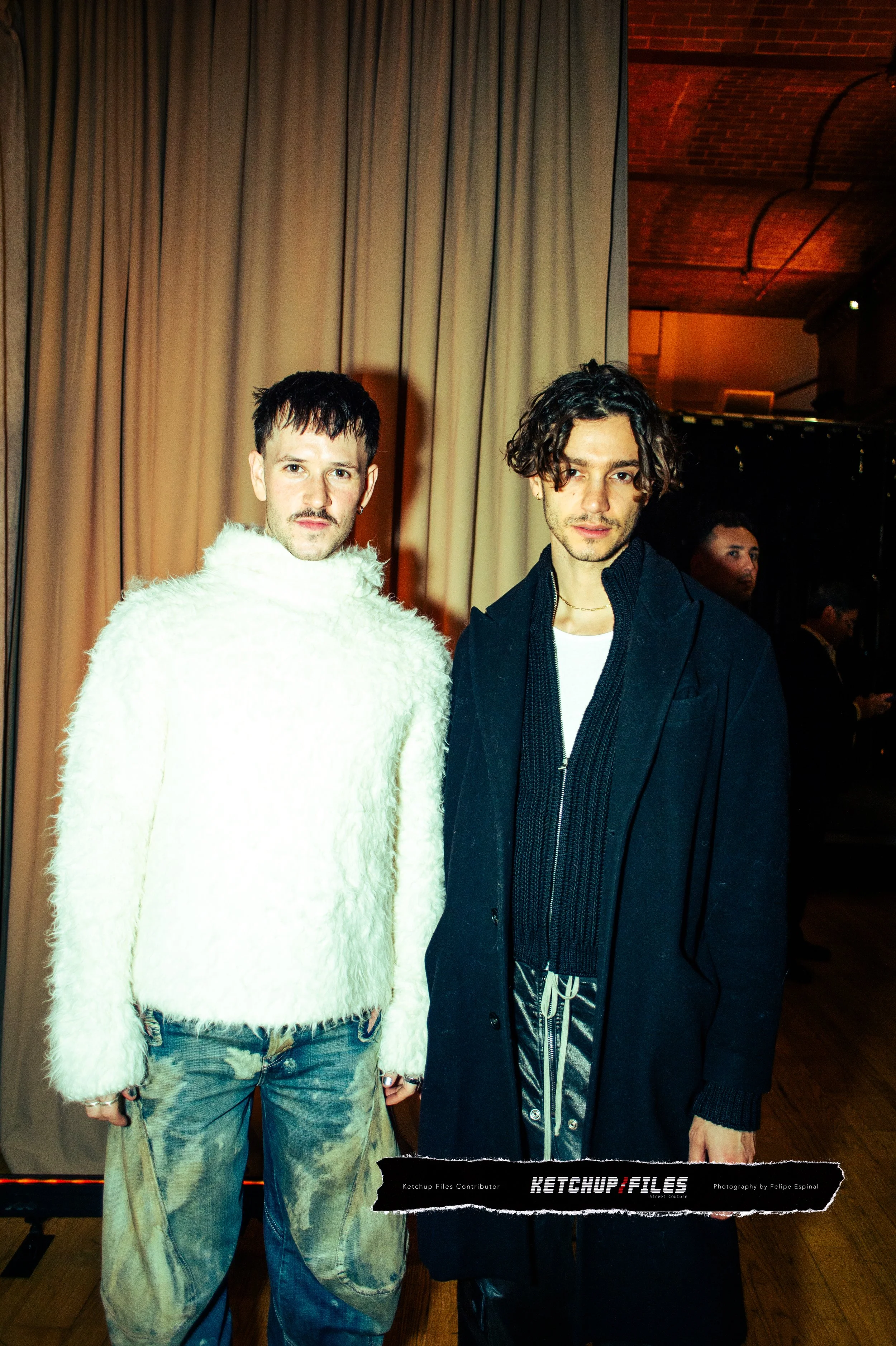 Two young men standing in front of a beige curtain at an indoor event, one wearing a white fuzzy sweater and jeans, the other in a black coat with a black sweater underneath.