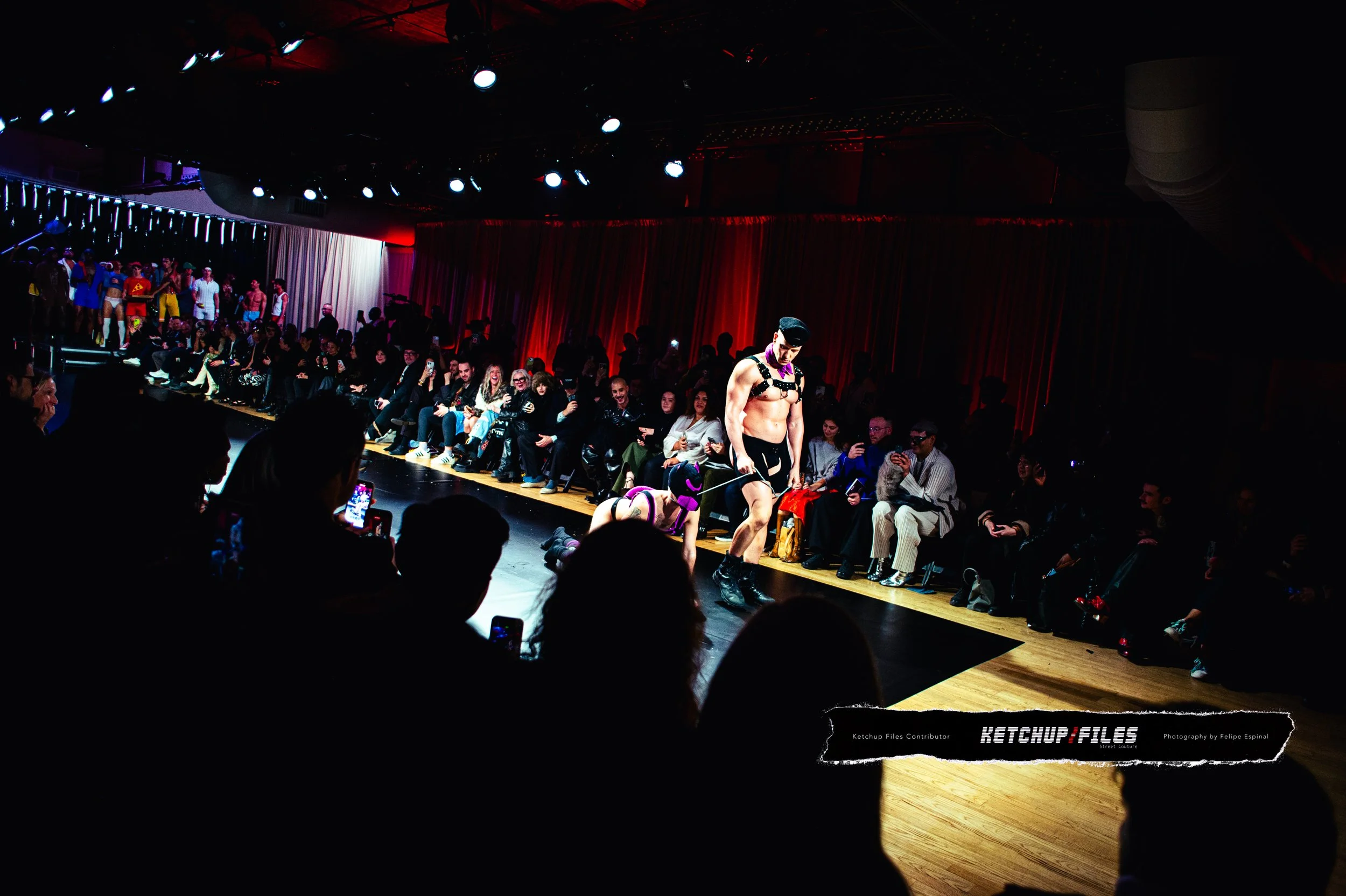A fashion runway show featuring a male model walking down the runway in an edgy, revealing outfit with bold accessories, while an audience watches from the seats and some take photos.