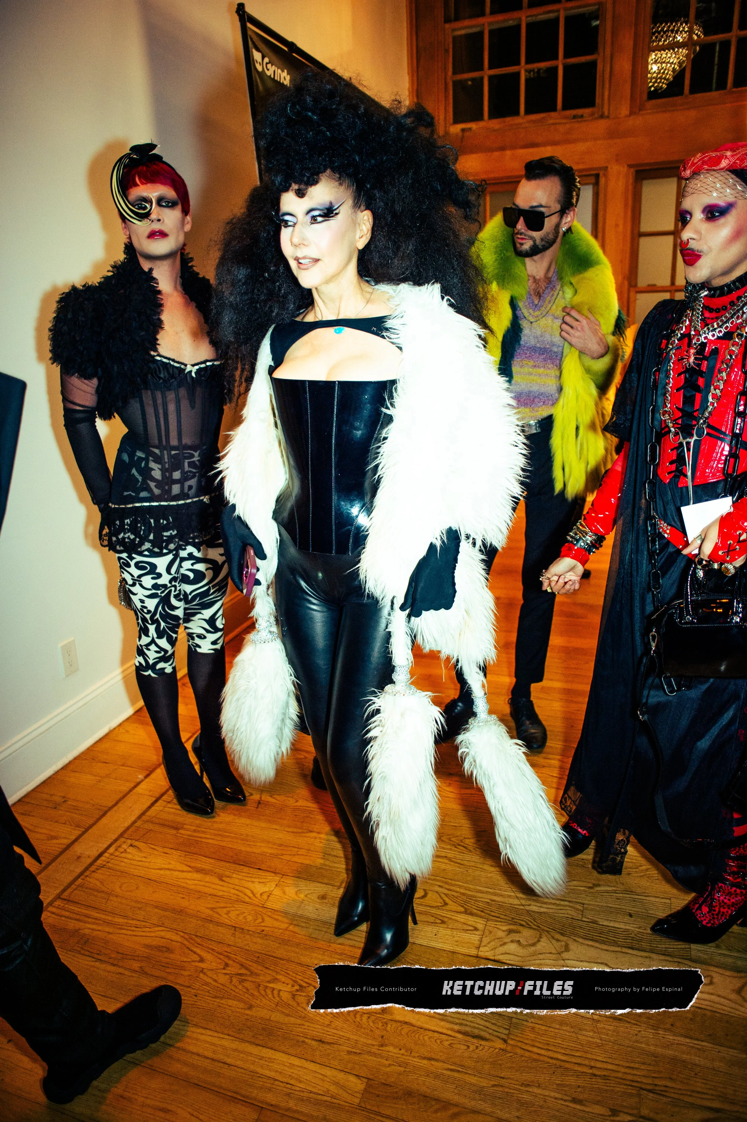 Group of people in colorful, eccentric outfits at a fashion event, with a woman in a black latex corset and ballet-length faux fur coat in the center.