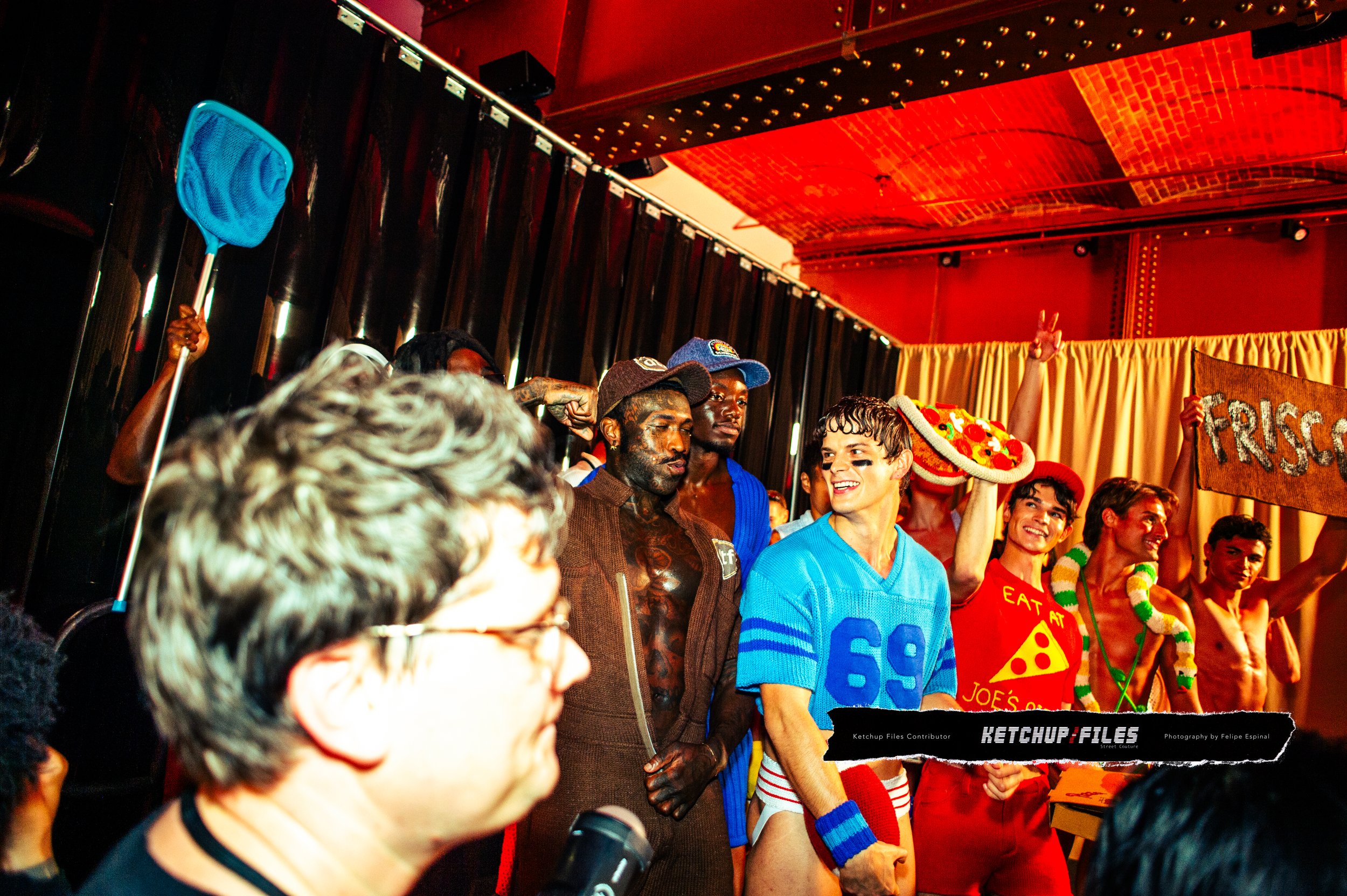 People dressed in colorful costumes on a stage, celebrating with a pizza slice hat and a sign that says 'FRE$O' in the background.