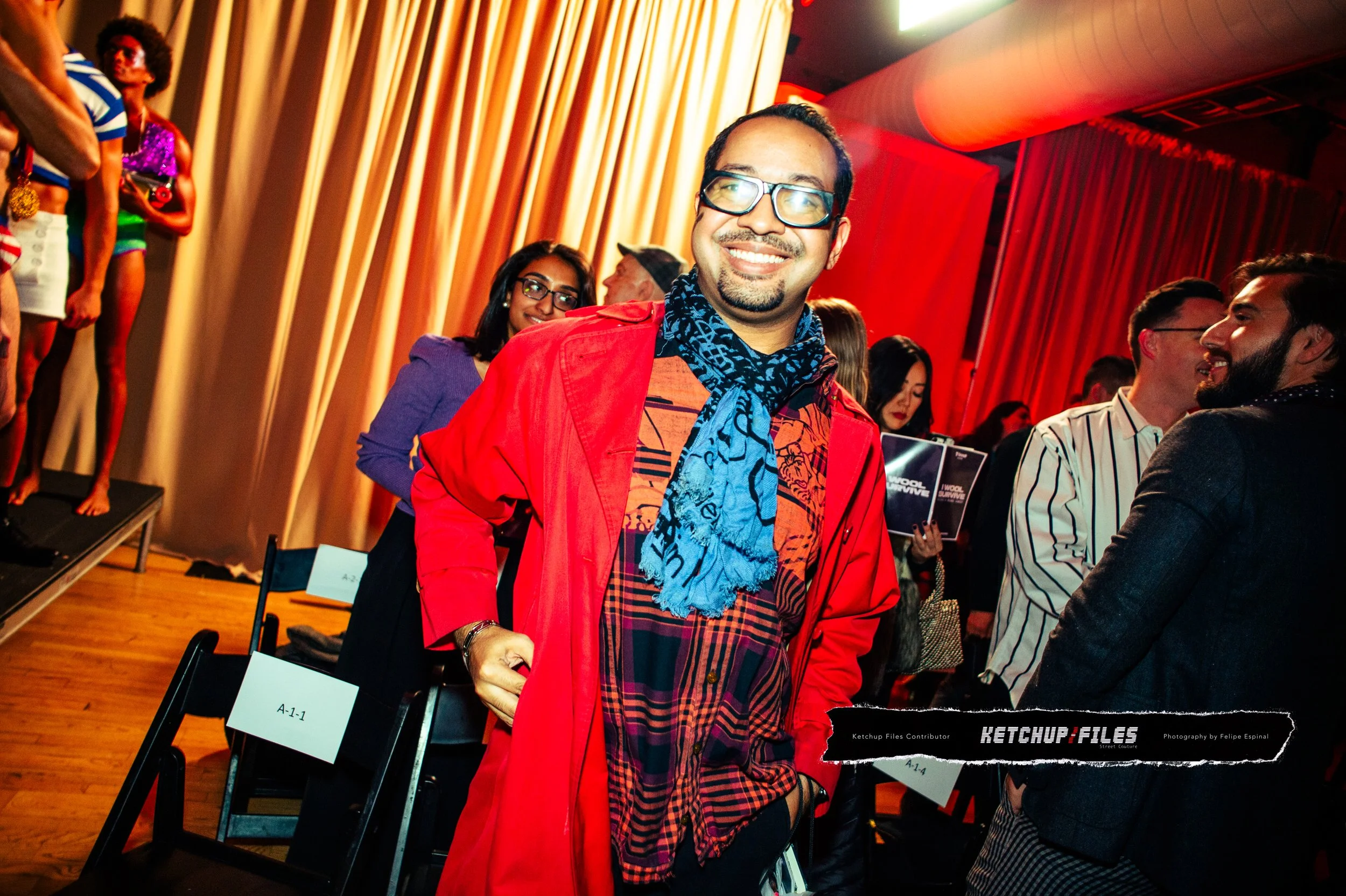 Smiling man in red jacket and glasses standing among people at an indoor event, with some holding books and others engaging in conversation.