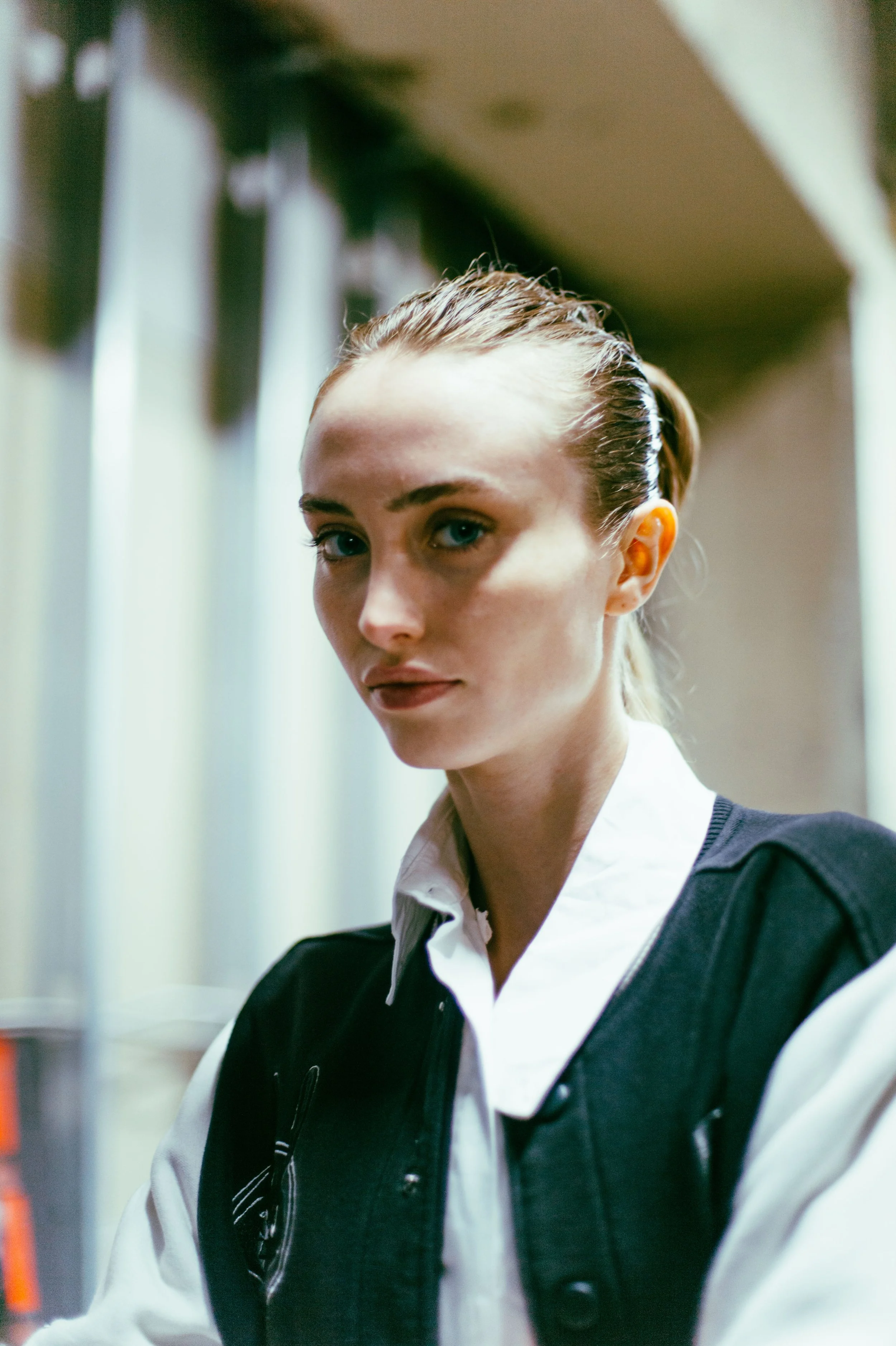Person with slicked-back hair, wearing a white shirt and dark jacket, standing indoors.