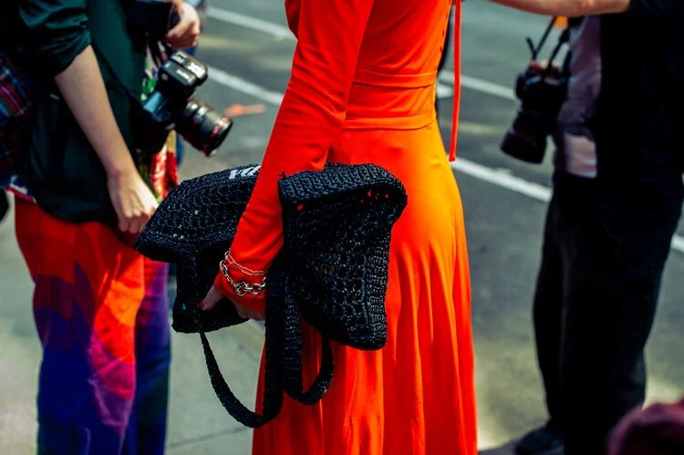 A woman in a bright orange dress holding a black woven bag, surrounded by photographers taking pictures.