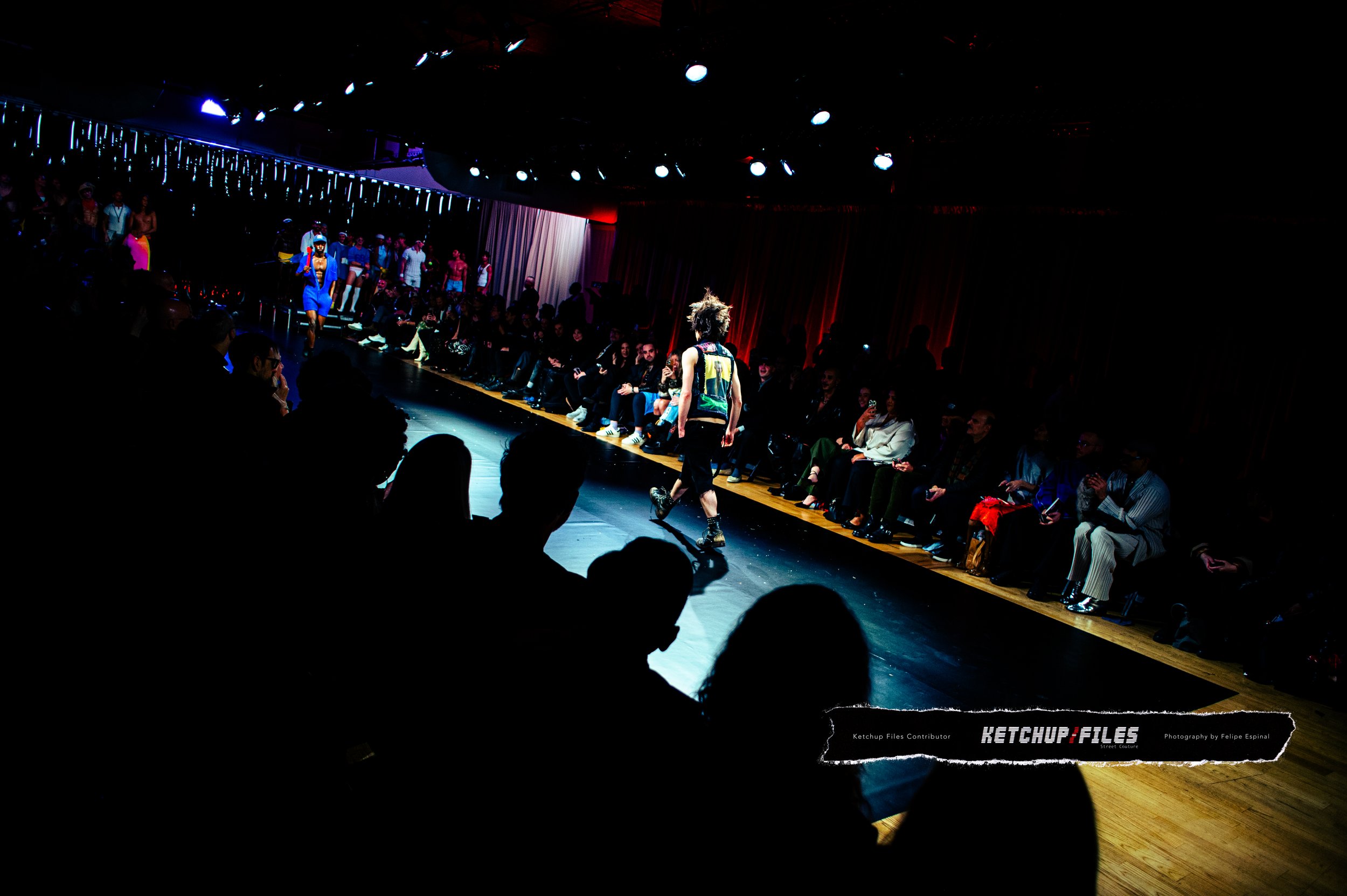 A runway fashion show with models walking down the catwalk surrounded by seated spectators, with colorful lighting and curtains in the background.