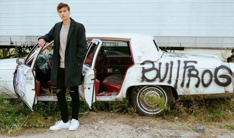 A young man stands next to an old, abandoned car with the word 'BULLROG' spray-painted on its side in black. The car's doors are open, exposing the interior, and it is parked on overgrown grass next to a light blue building.