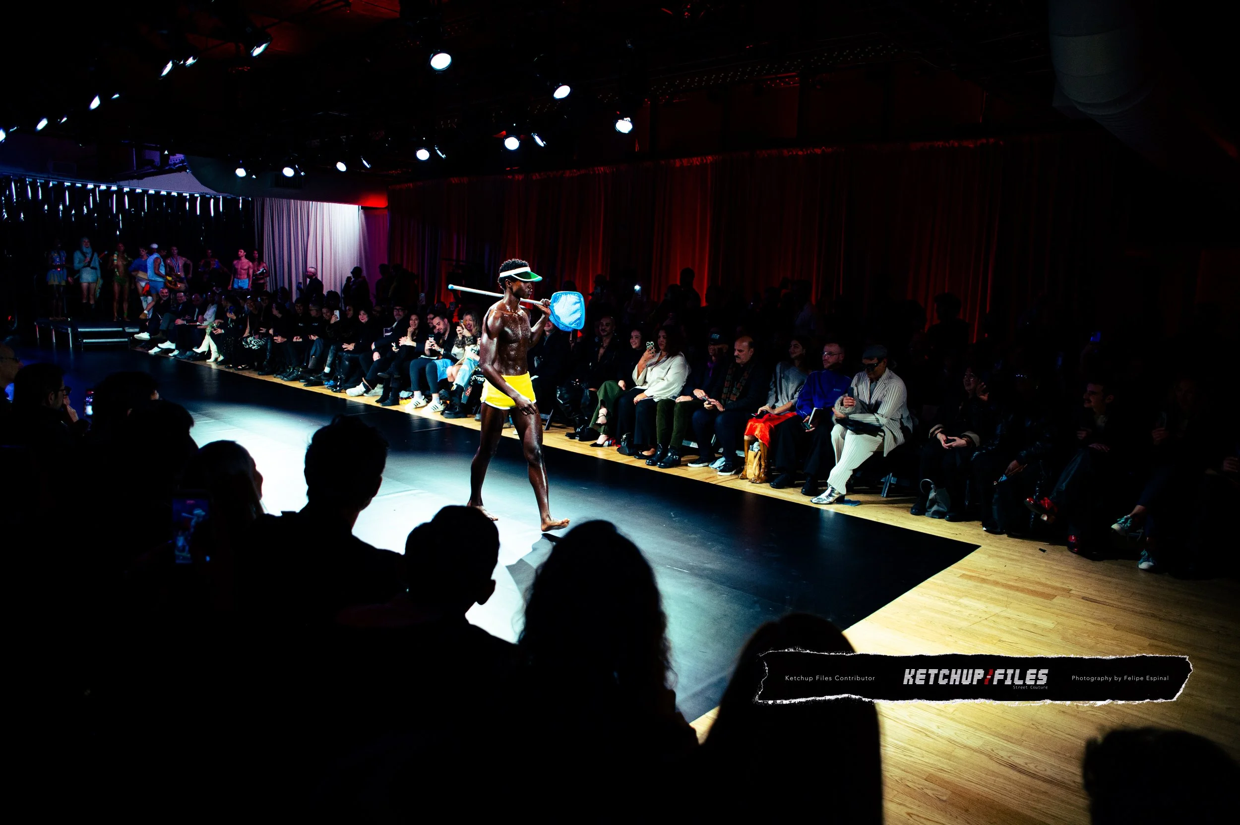A person walking on a runway at a fashion show, wearing yellow shorts, a visor, sunglasses, and holding a net. Audience members sit along the runway watching the event.