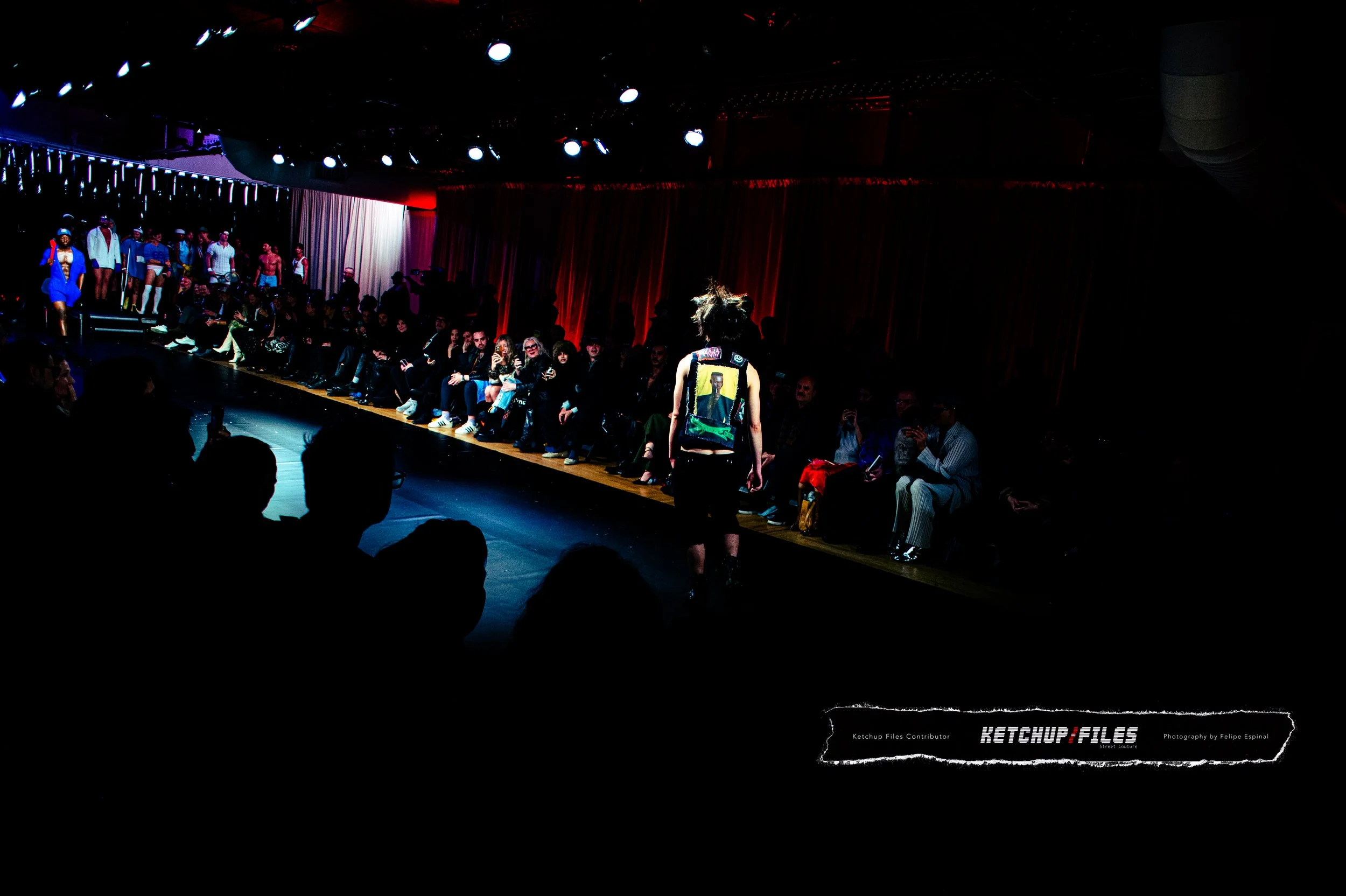 A fashion runway show with a model walking on a dark runway and an audience seated on both sides, with colorful lighting and a red curtain backdrop.