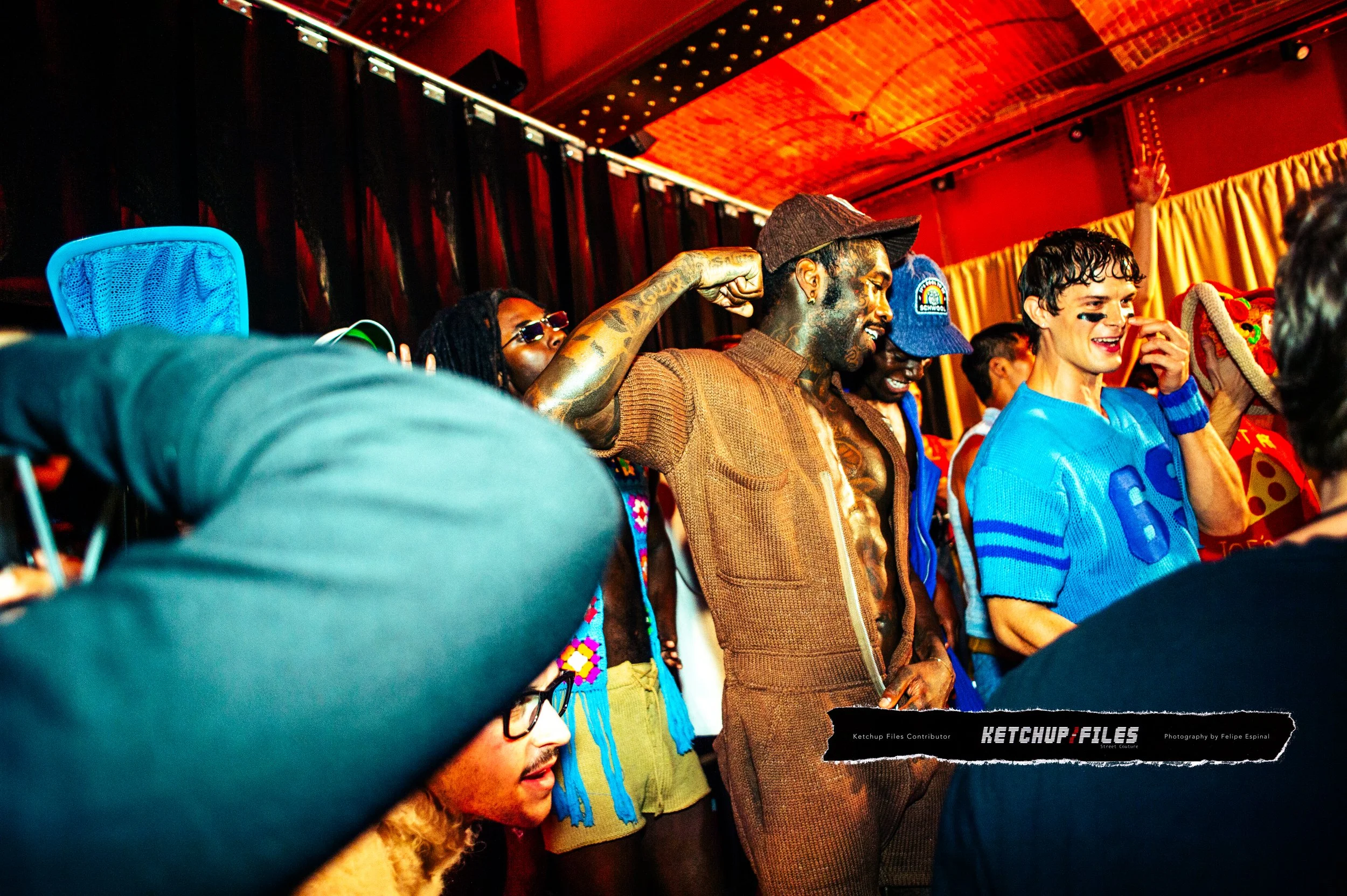 Group of people at a party or event, some smiling and others posing, with a man in the center flexing his arm. The setting appears indoors with red walls and curtains.