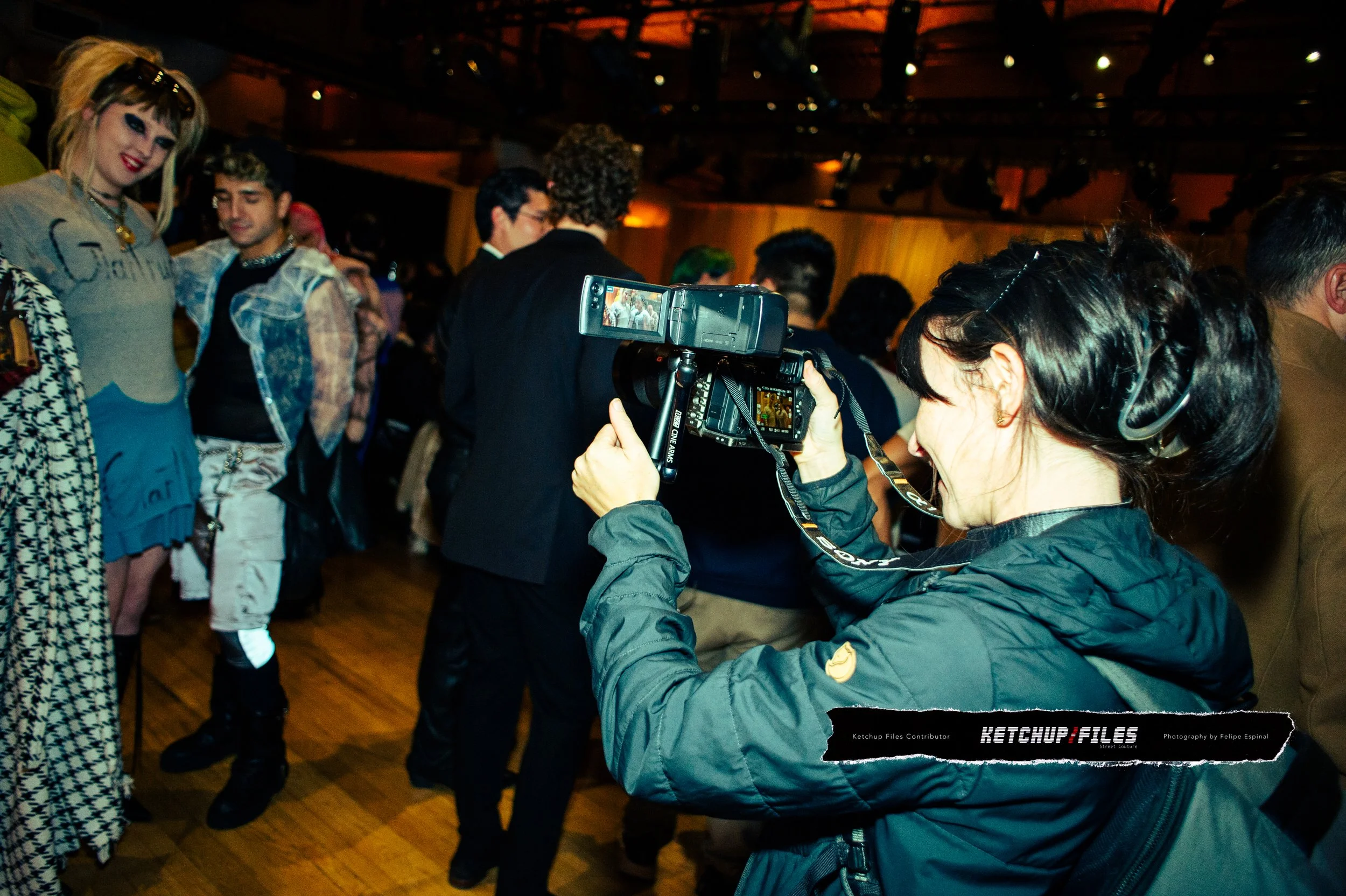 A woman with dark hair holding a camera, taking a photo at a lively indoor event with many people in the background, some posing and others talking.