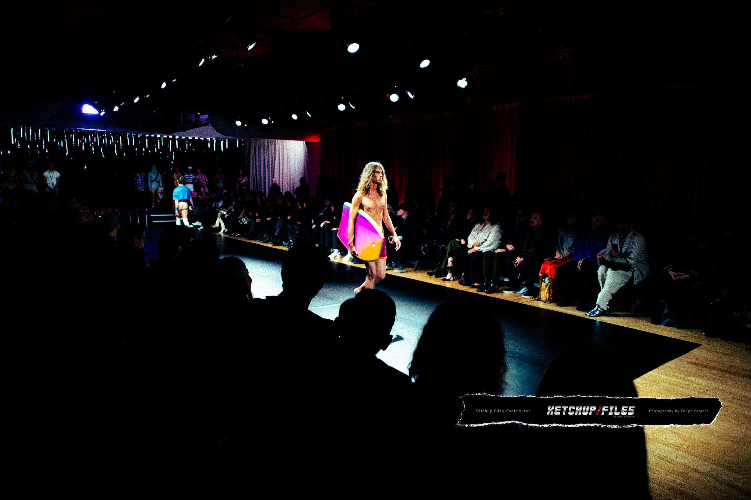 A fashion runway show with a male model walking down the runway carrying a large pink and yellow object, possibly a surfboard, with audience seated on both sides and some models in the background.