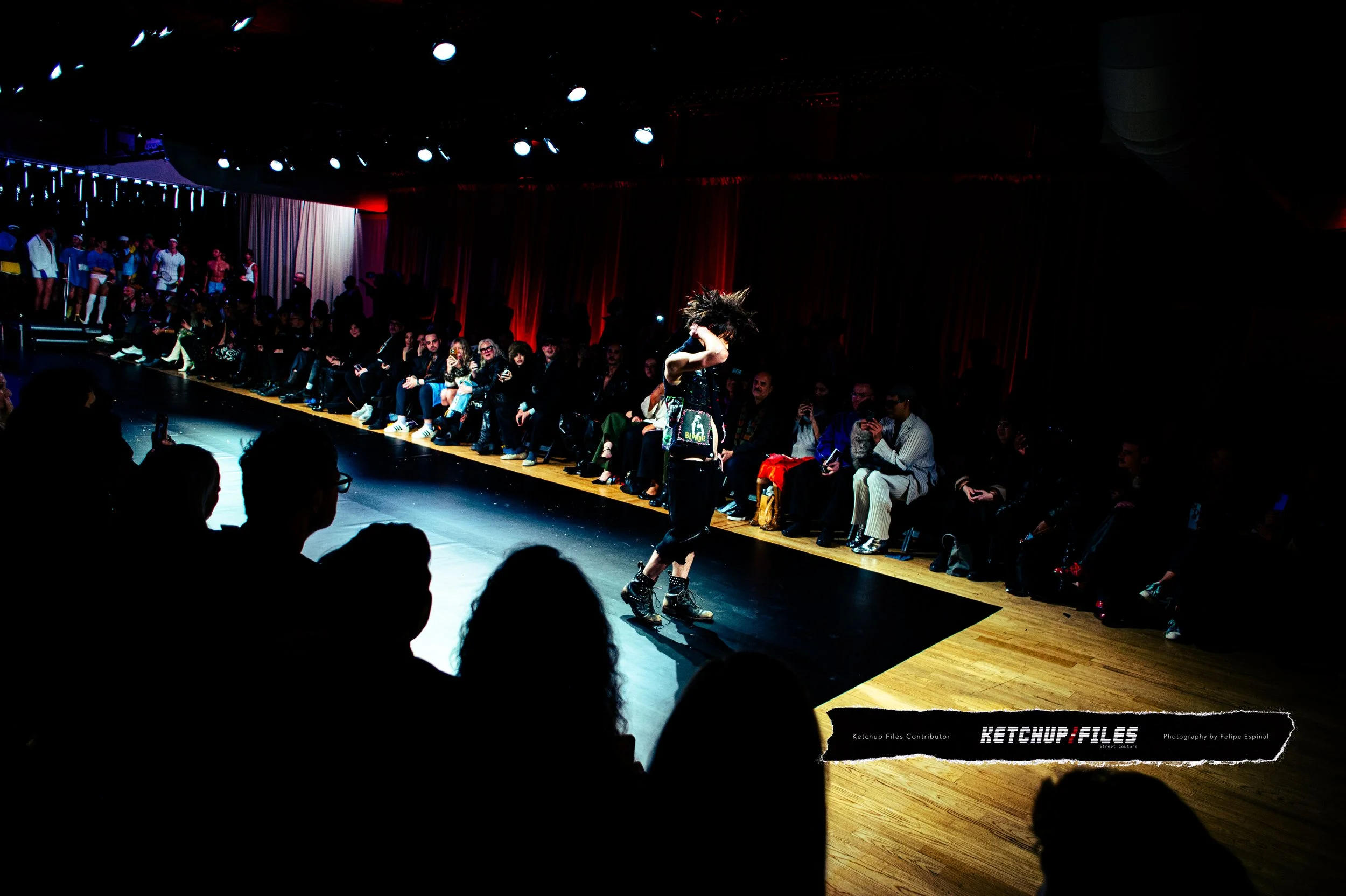 A performer dancing on a lit stage during a fashion or dance event with an audience sitting around the stage in a darkened room.