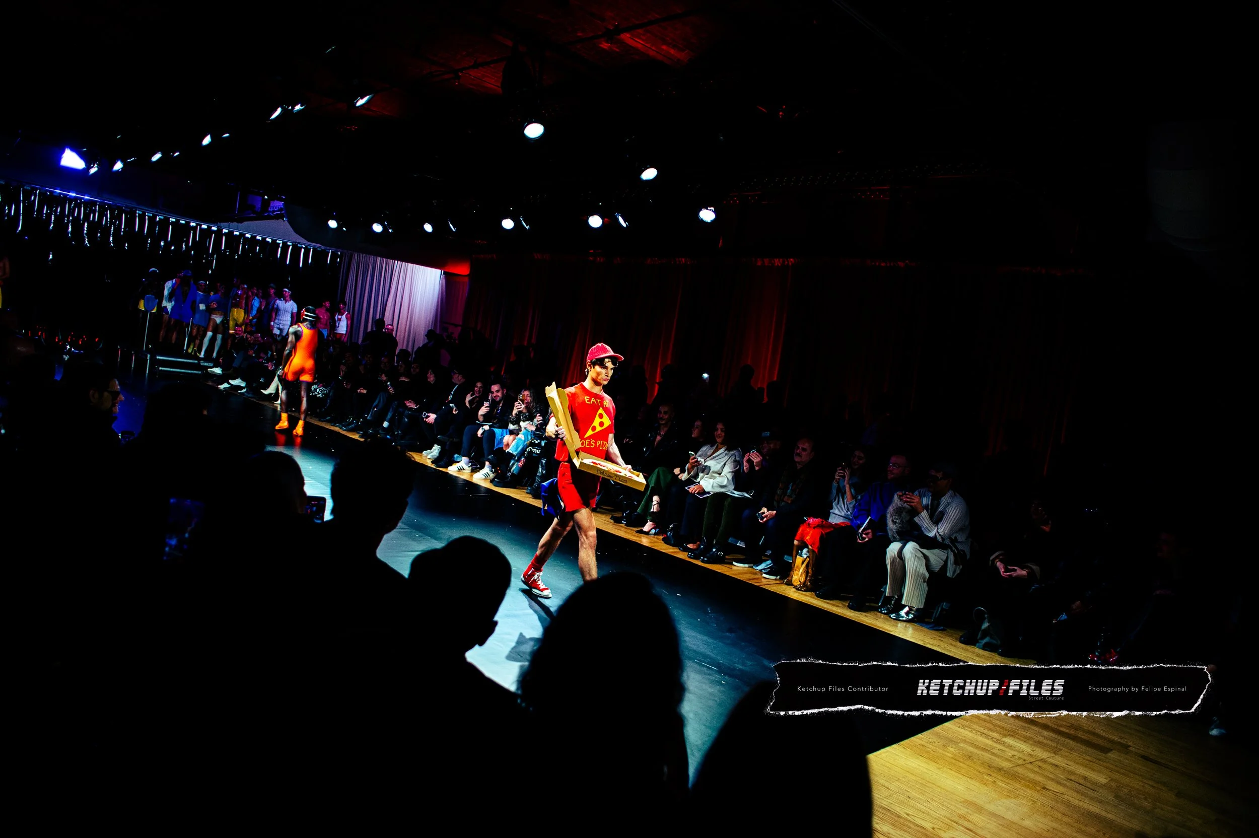 Fashion runway show with a model dressed as a pizza delivery person carrying a pizza box on the runway, audience seated on both sides, dark lighting with spotlights.