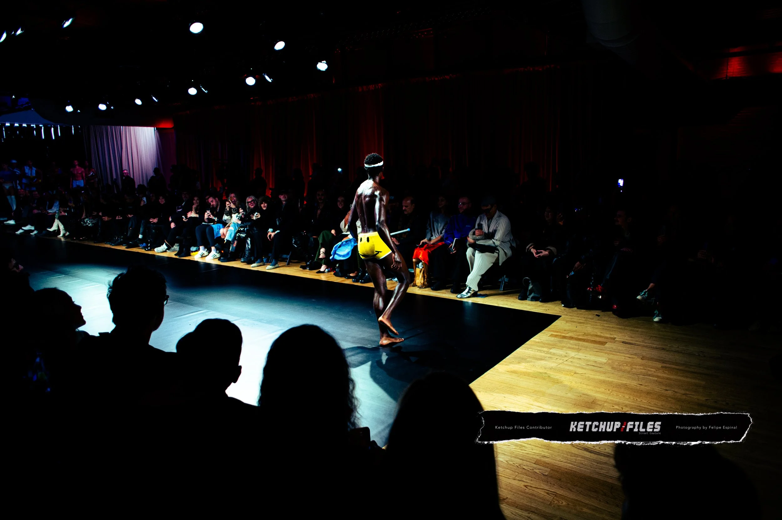 A performer on a runway at a fashion show, wearing bright yellow shorts and a headband, with an audience seated around watching.