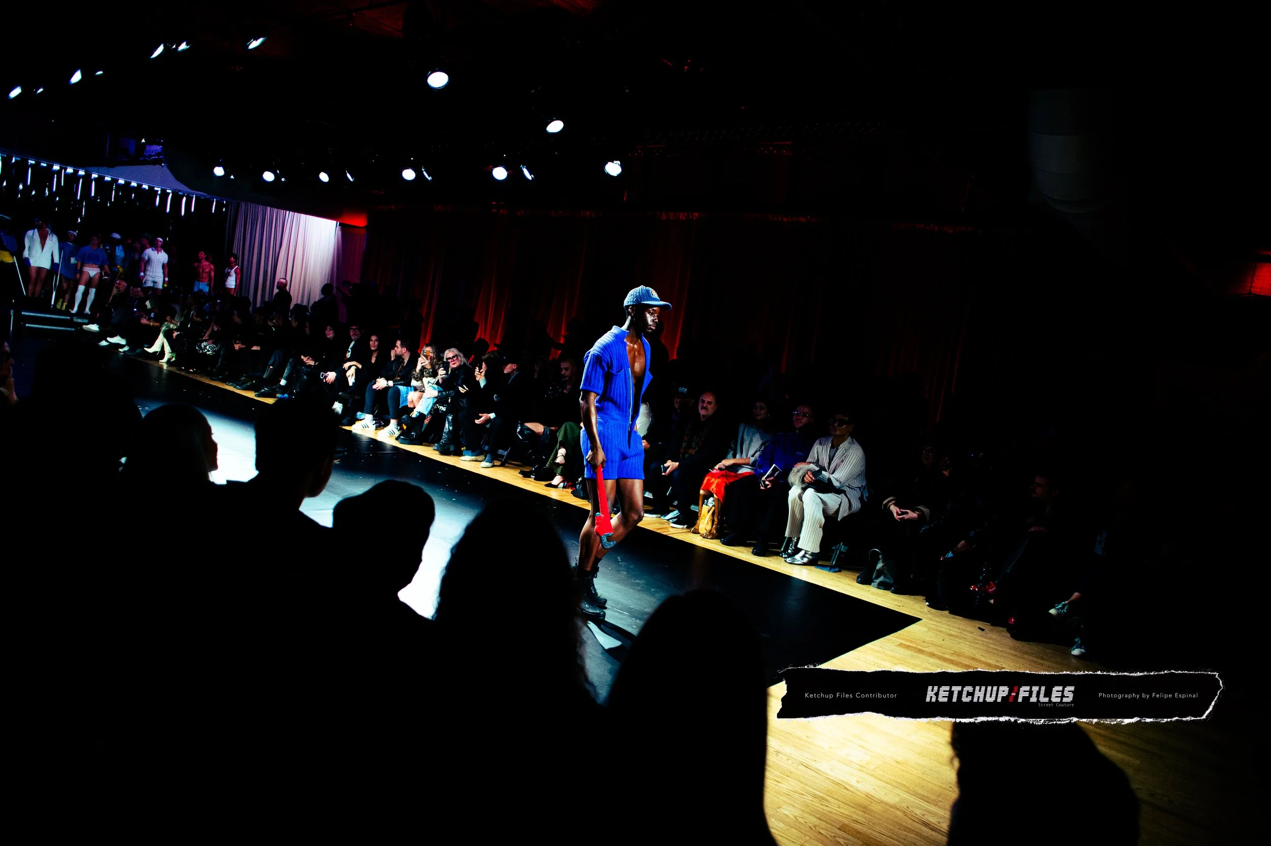 A male model walks on a runway during a fashion show, wearing a blue outfit and a cap, with an audience seated on both sides in a dimly lit room with red curtains in the background.