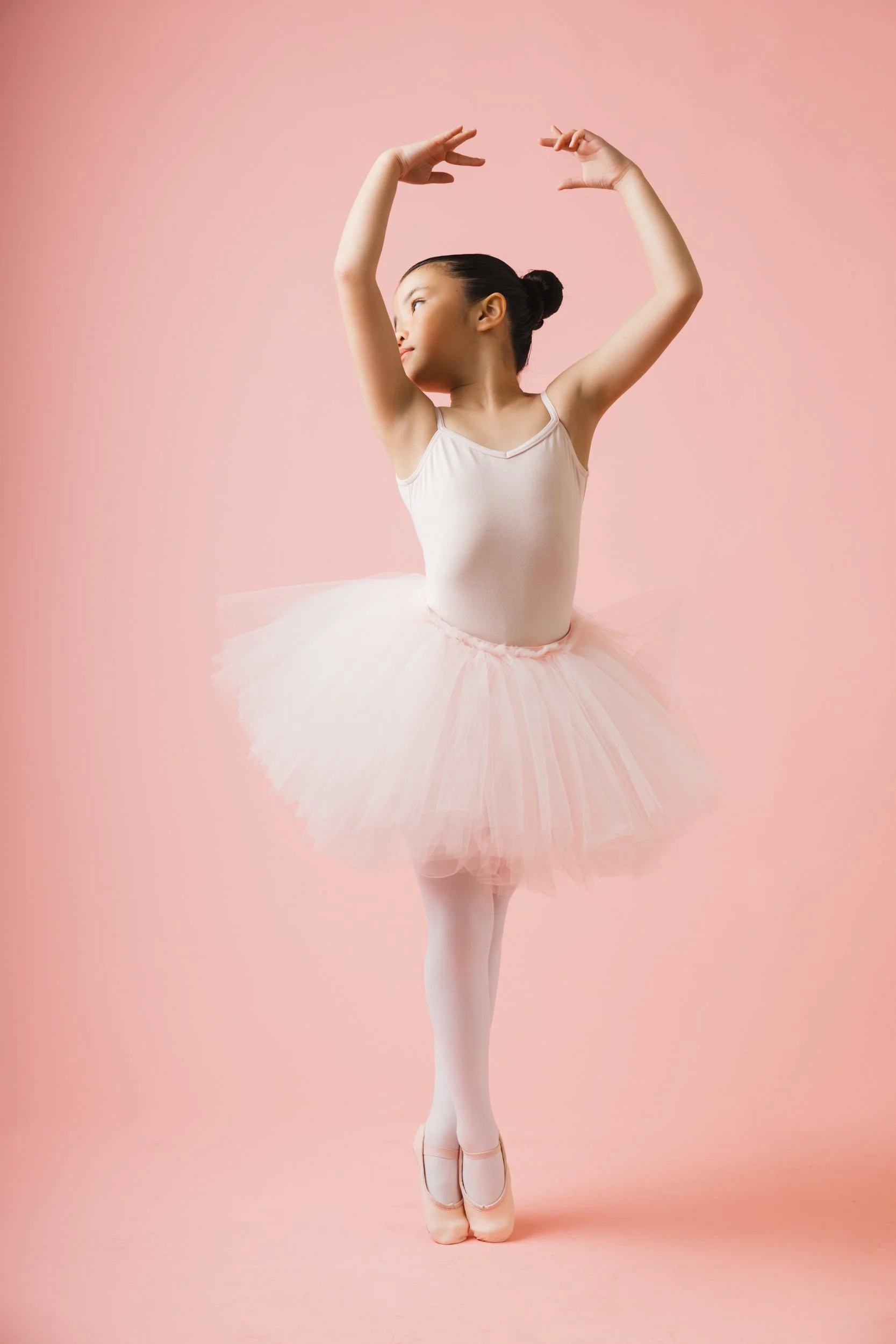 Young girl in ballet attire, wearing a light pink leotard, tulle tutu, tights, and pointe shoes, poses with arms raised and head turned against a pink background.