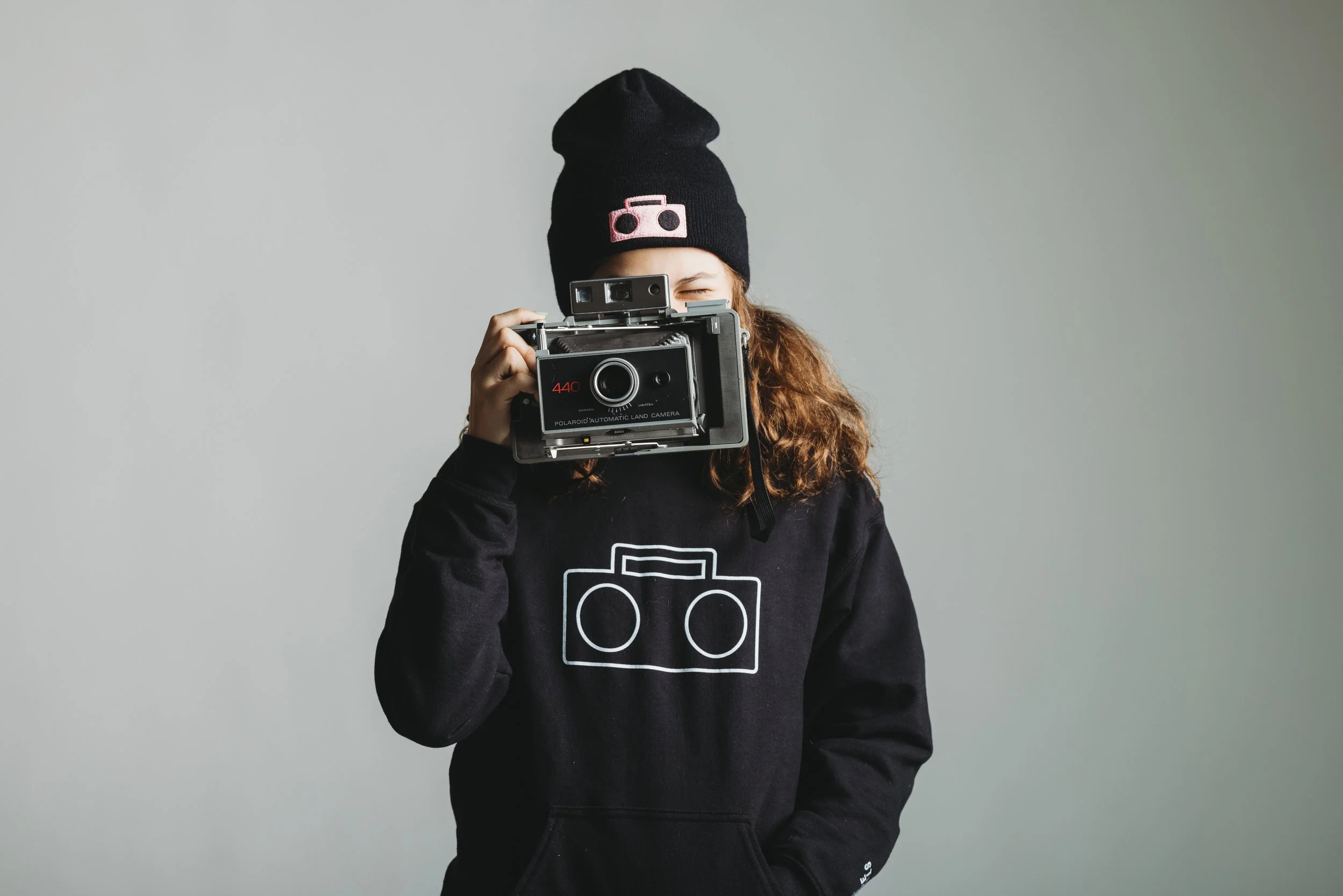 A young person with curly hair wearing a black beanie and black sweatshirt with a camera graphic, holding a vintage Polaroid camera up to their face and taking a photo.