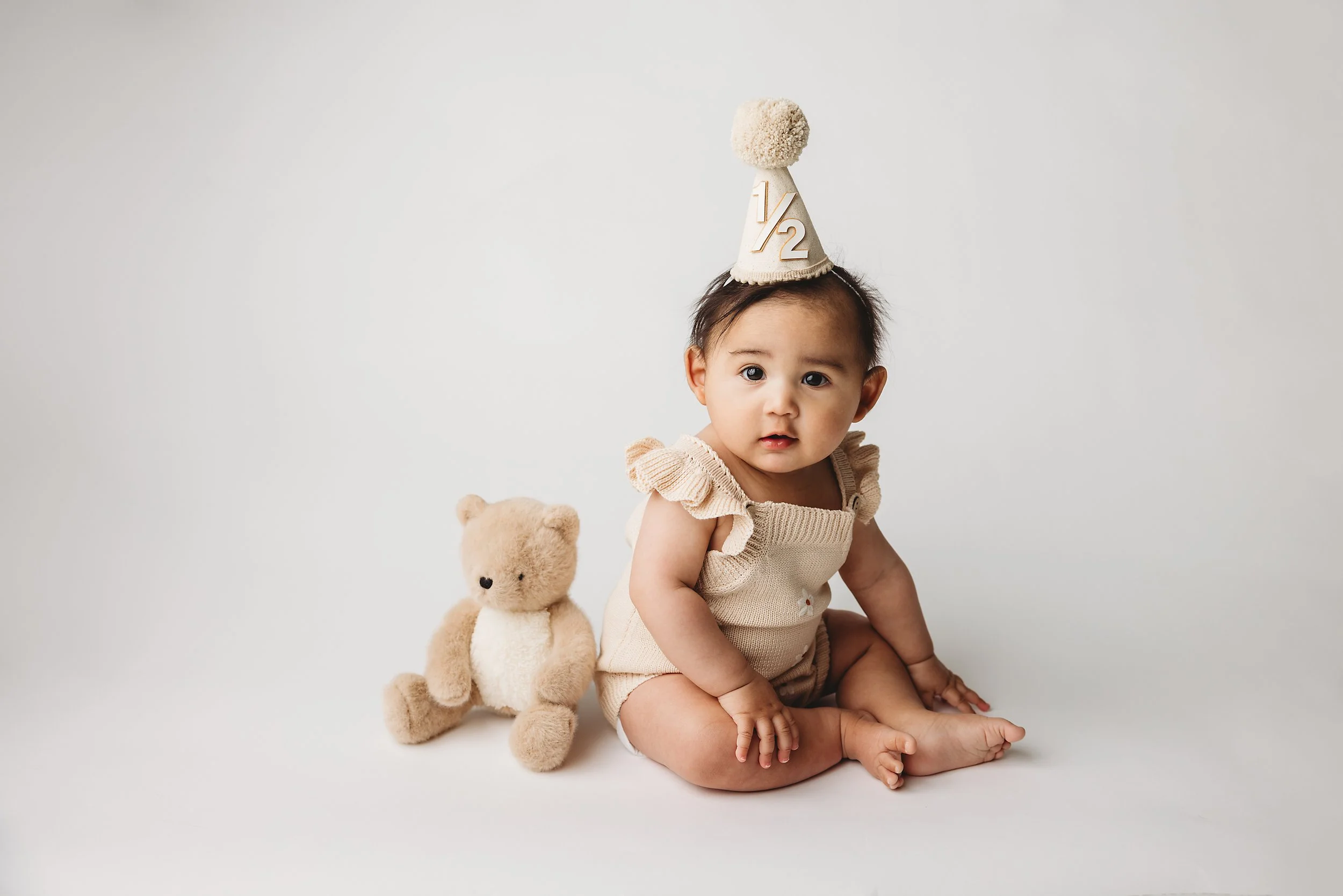 A baby sitting on the floor with a teddy bear, wearing a cream-colored outfit and a birthday hat with '1/2' on it.