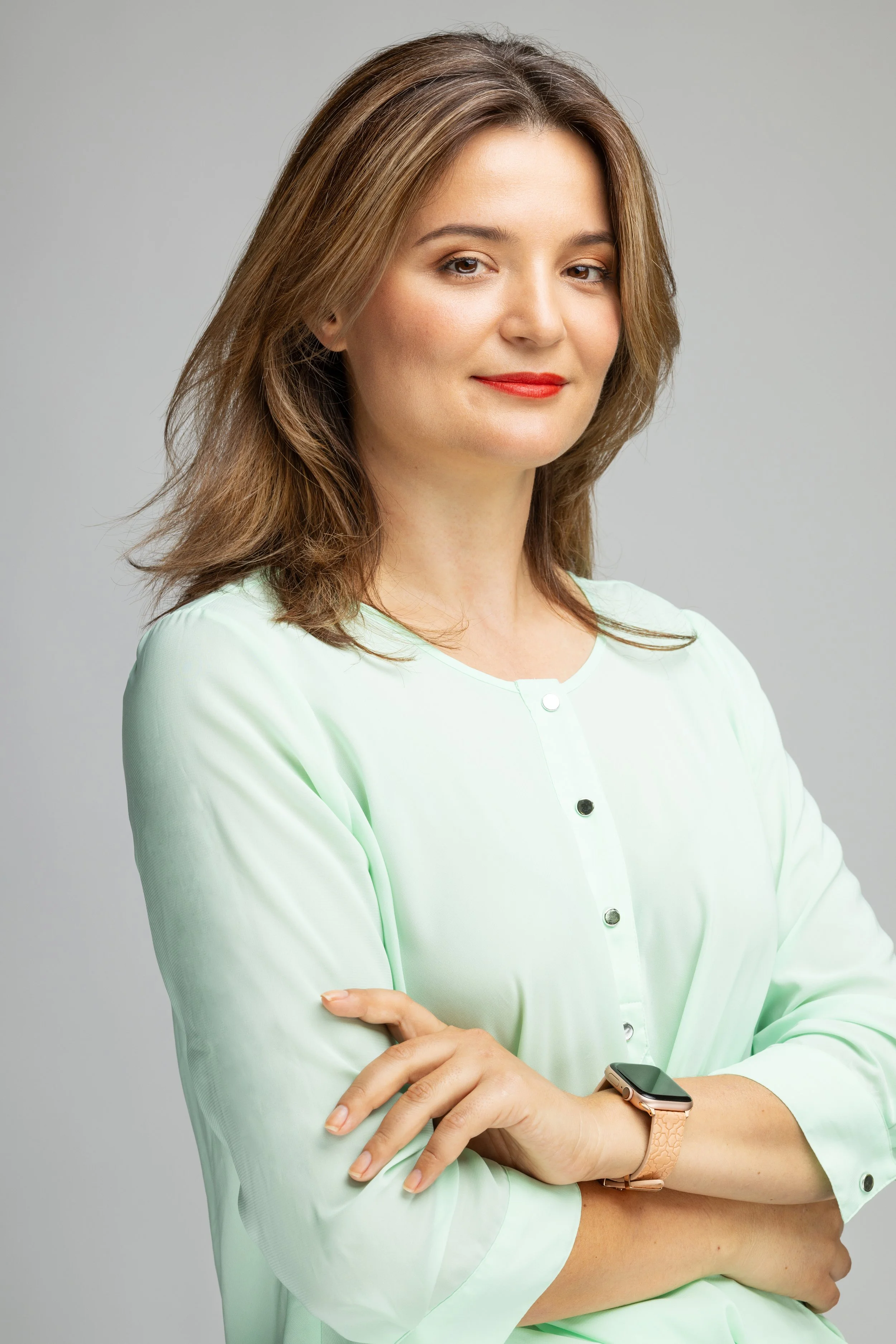 A woman with shoulder-length brown hair, wearing a light green blouse, red lipstick, and a smartwatch, standing with her arms crossed against a plain grey background.