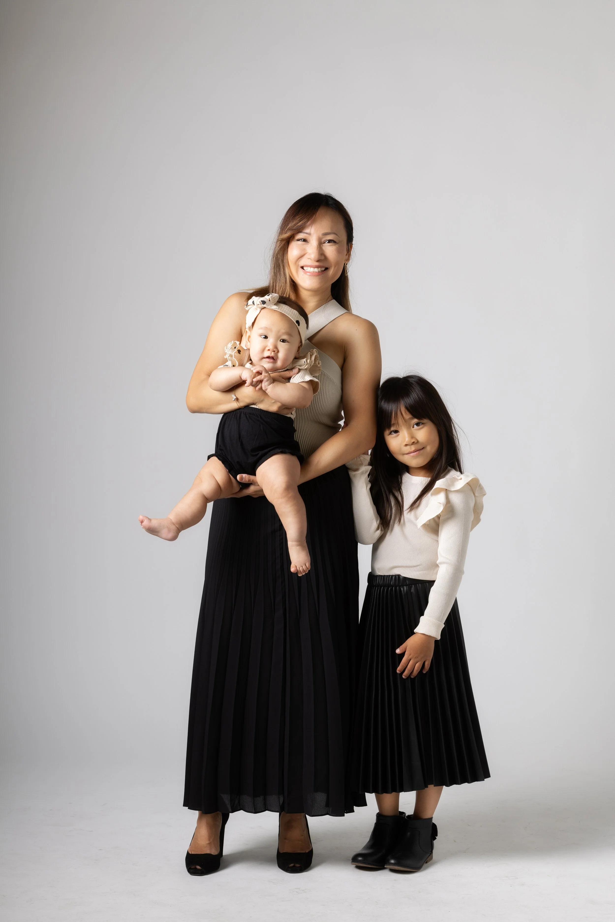 A woman smiling and holding a baby girl, with an older girl standing next to her, all dressed in stylish outfits against a plain background.