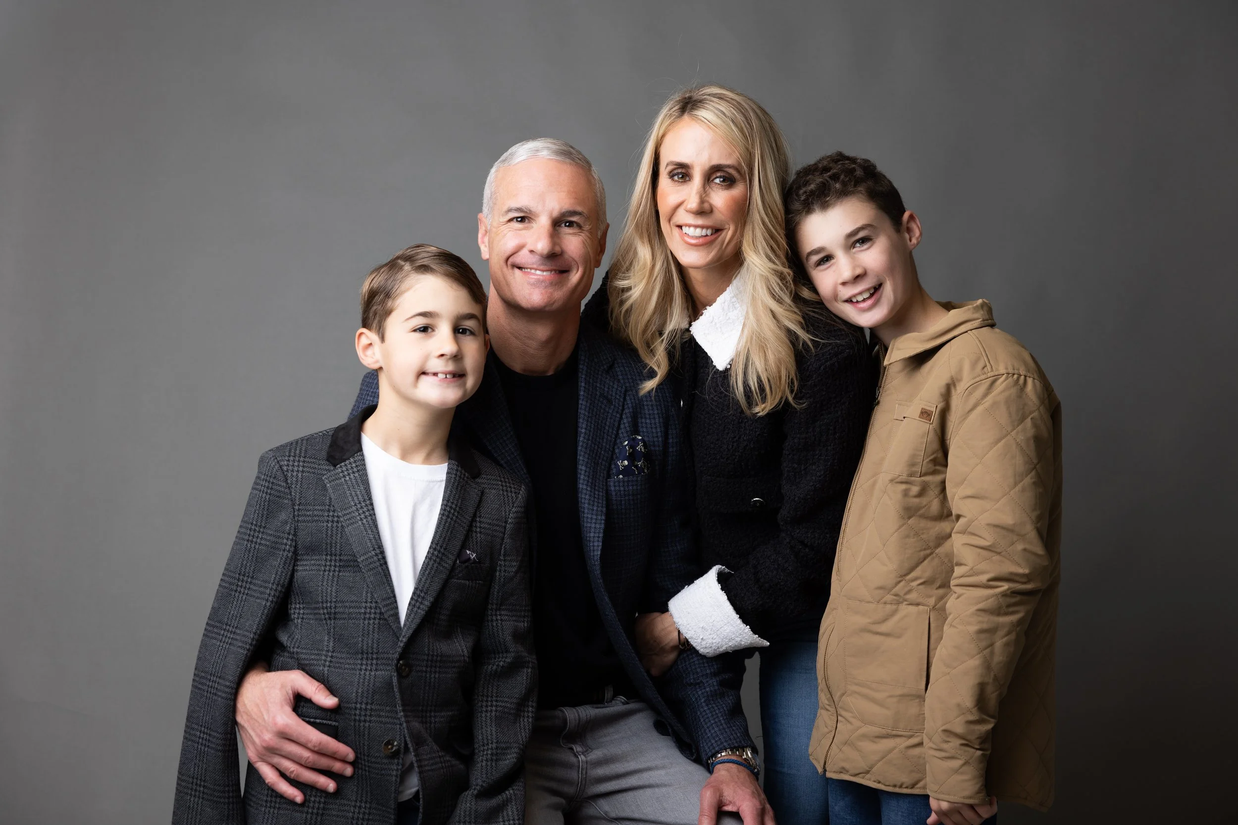 A family of five posing together against a plain gray background. The family includes two children, a man, a woman, and an older man, all smiling and close to each other.