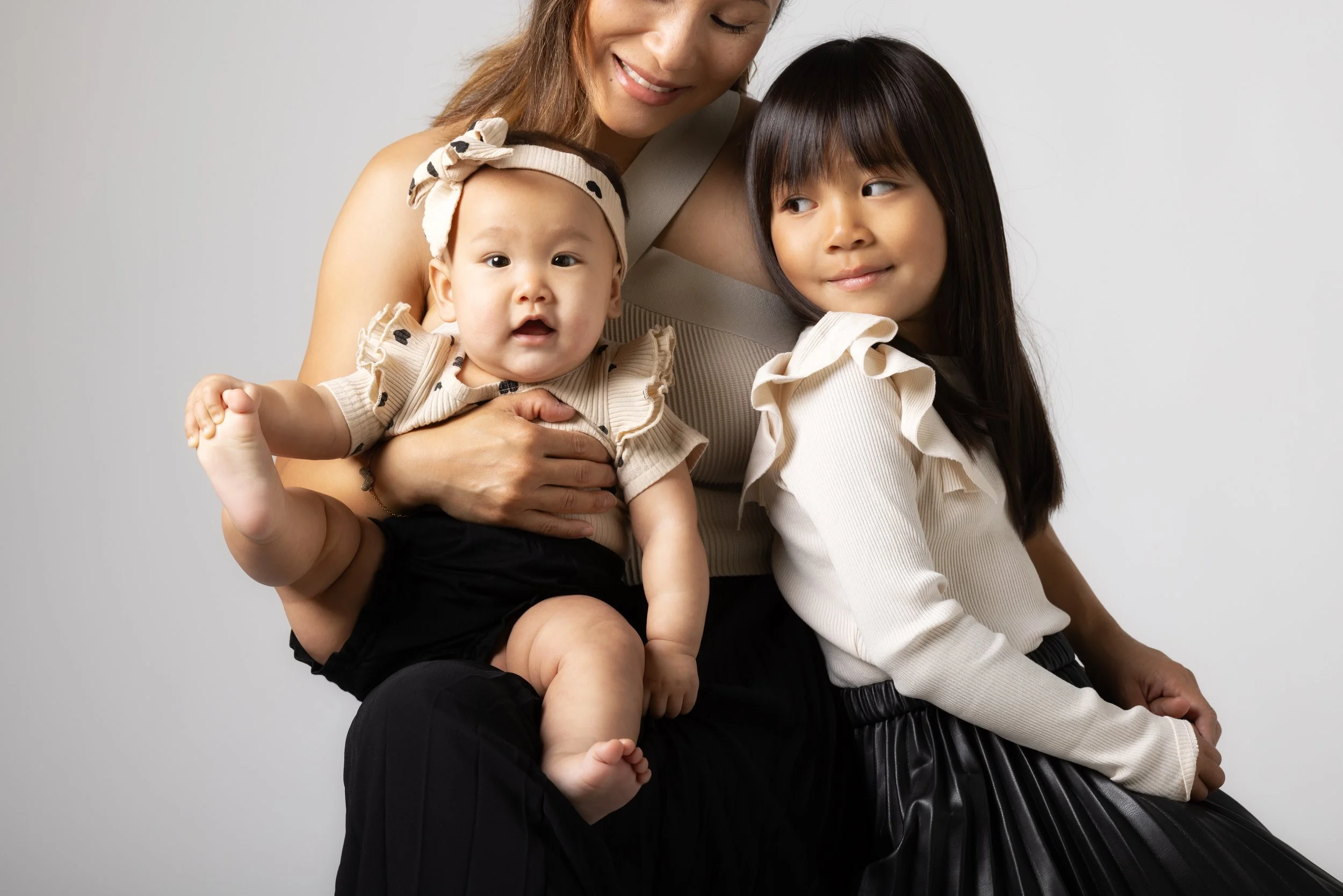 A woman with shoulder-length brown hair holding a baby girl with a beige headband and a beige shirt with black polka dots, and a young girl with long dark hair and a light-colored top, all smiling and sitting against a plain white background.