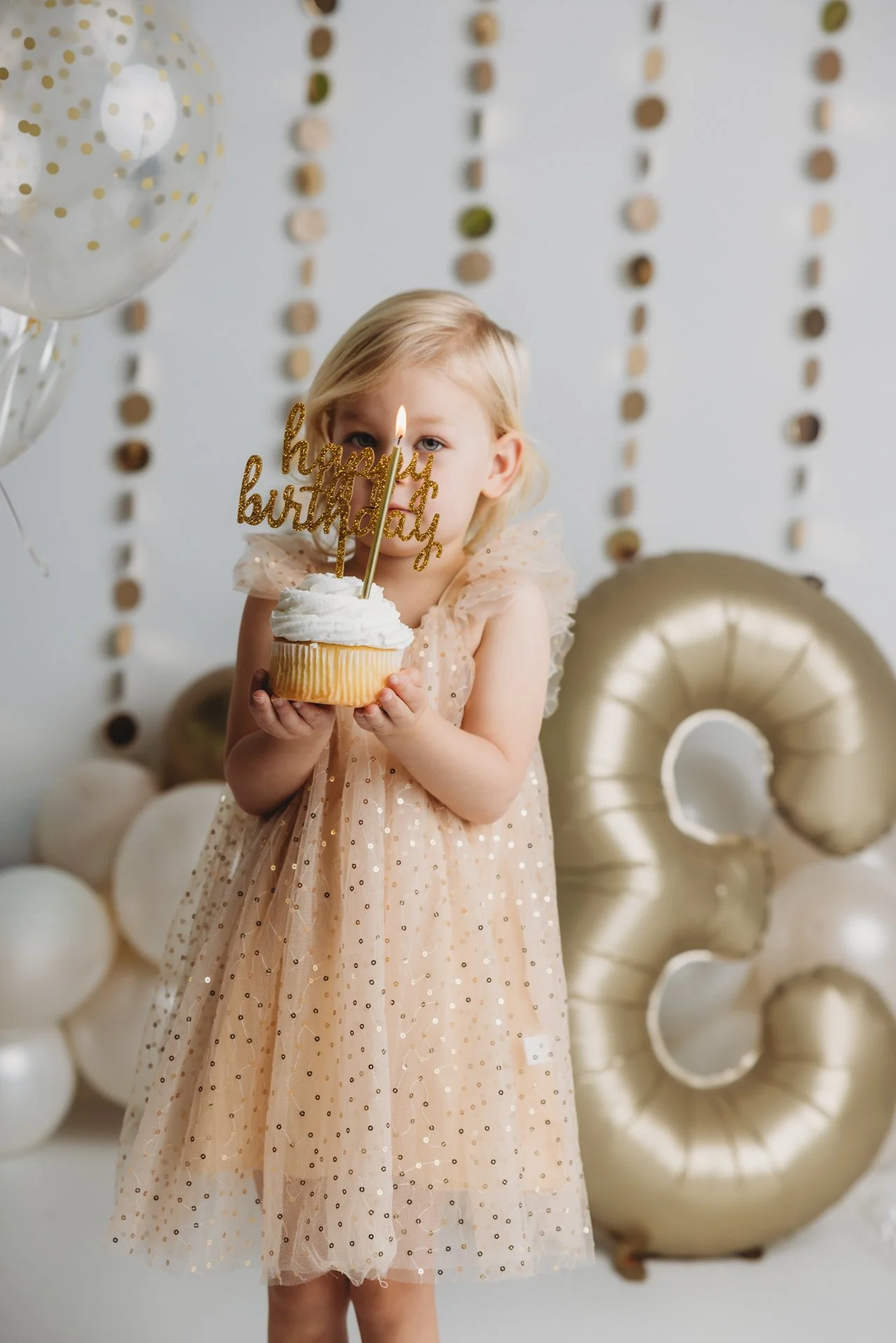 3 year old girl holding a cupcake with a birthday candle and sign in studio with balloons