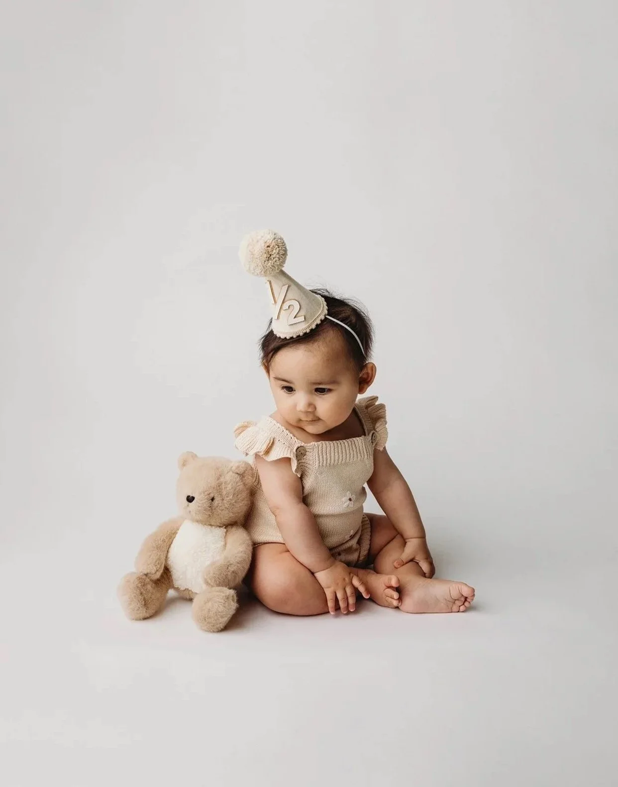 A young child sitting on the floor wearing a birthday party hat, with a teddy bear next to them, celebrating their second birthday.
