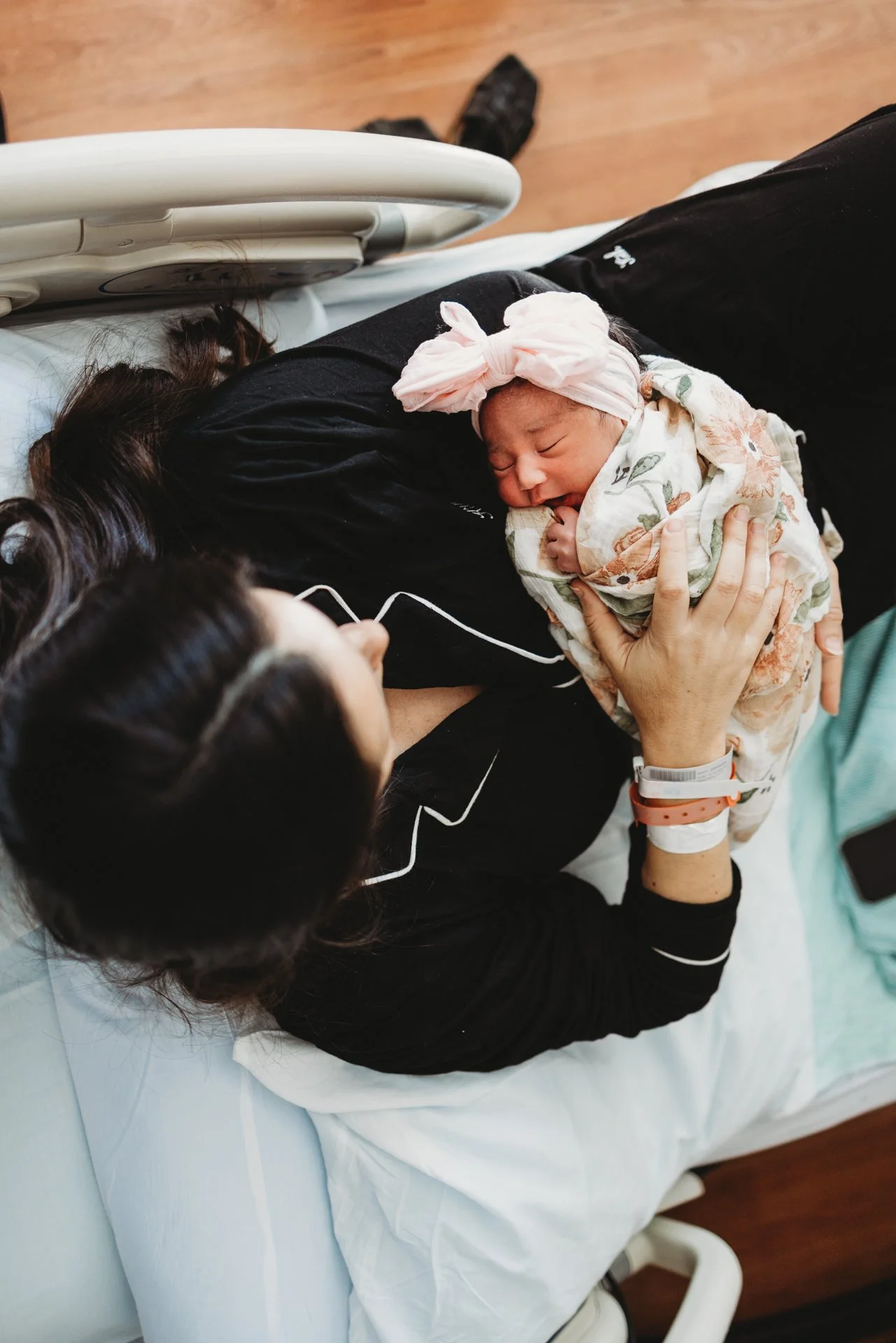 A newborn baby wrapped in a floral blanket with a pink headband sleeping on a woman's chest during childbirth in a hospital room.