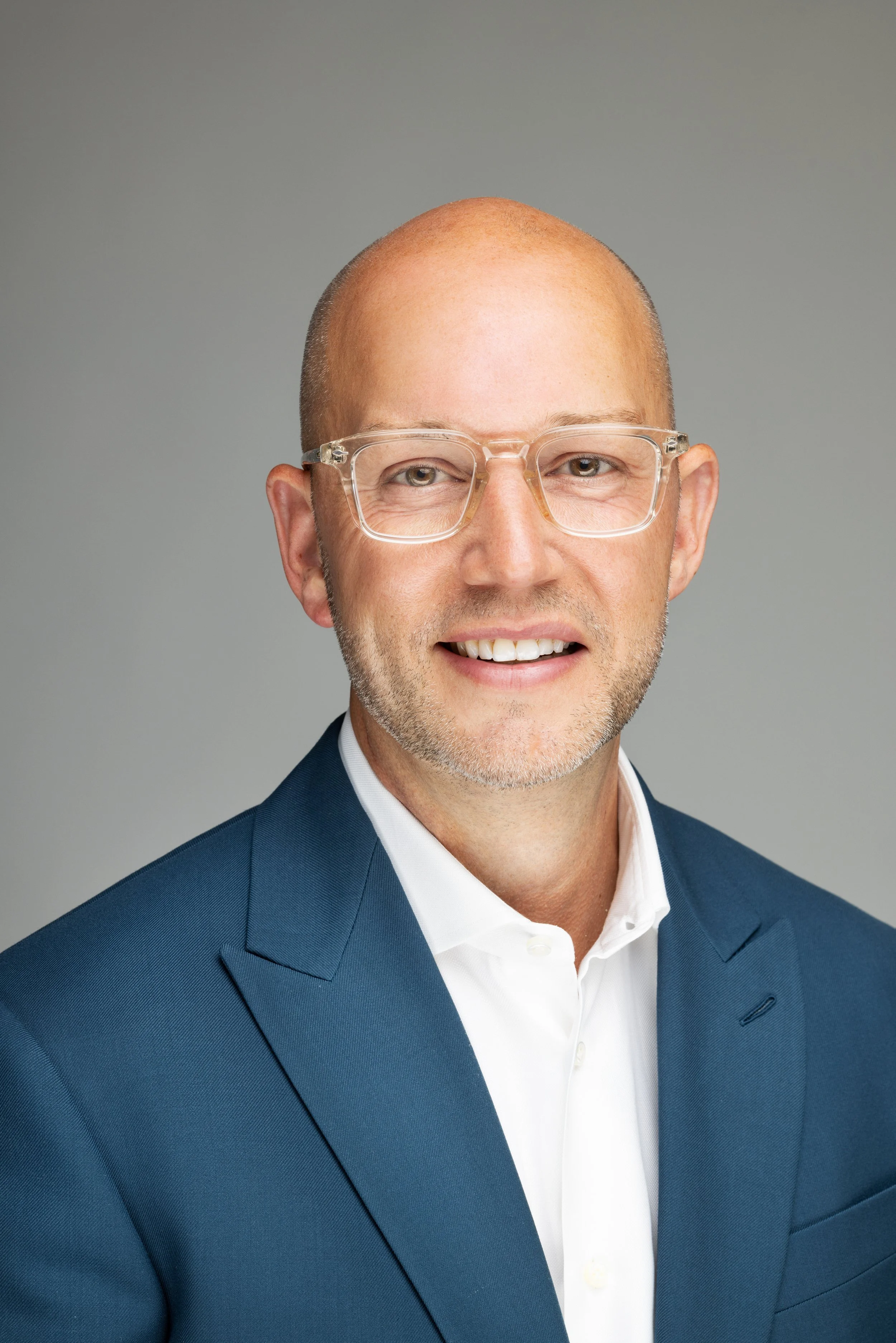 Headshot of a man with glasses, bald with some facial hair, wearing a blue suit and white shirt, smiling against a gray background.