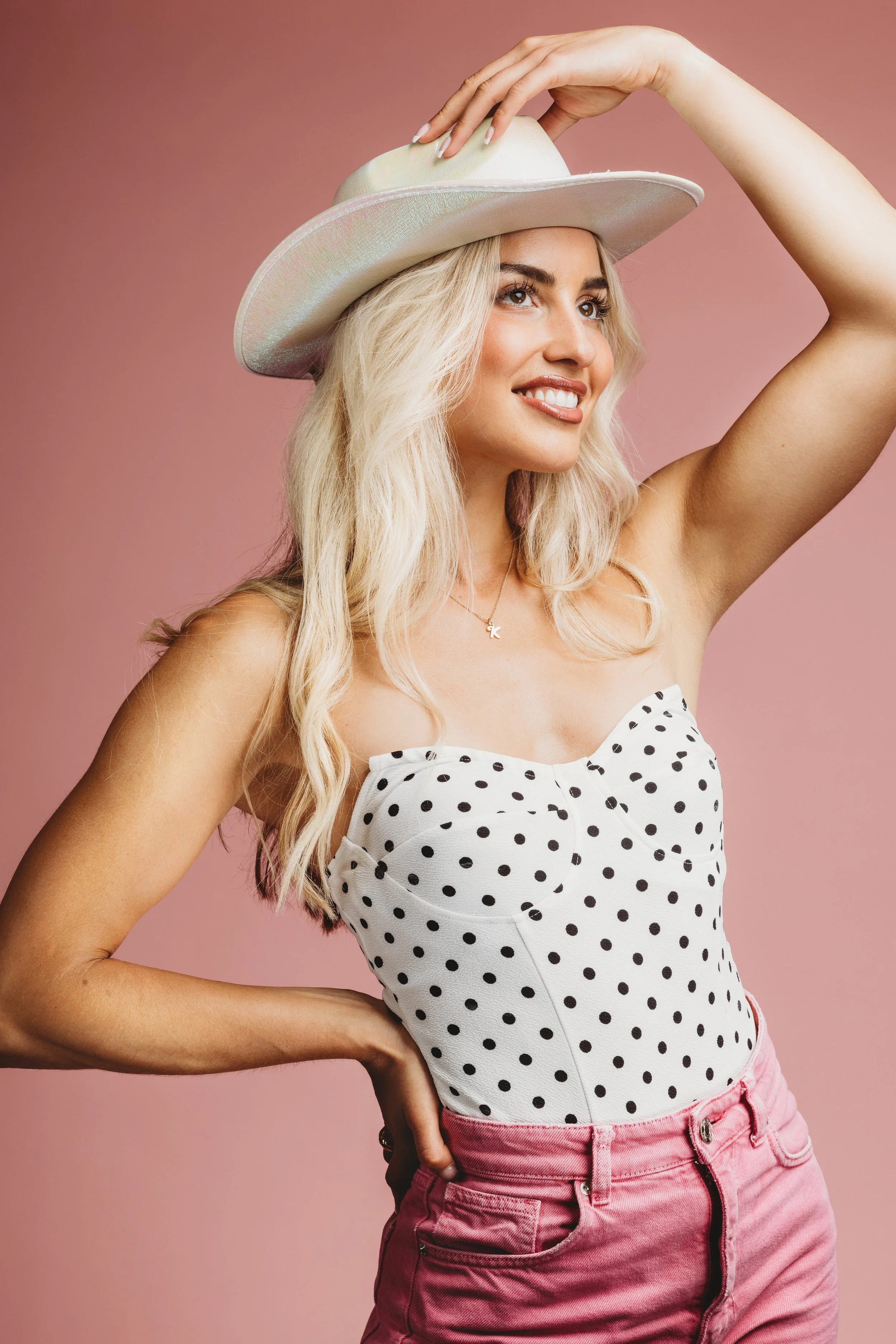 A young woman with long blonde hair wearing a white hat, a polka dot strapless top, pink pants, and a necklace, smiling and posing against a pink background.