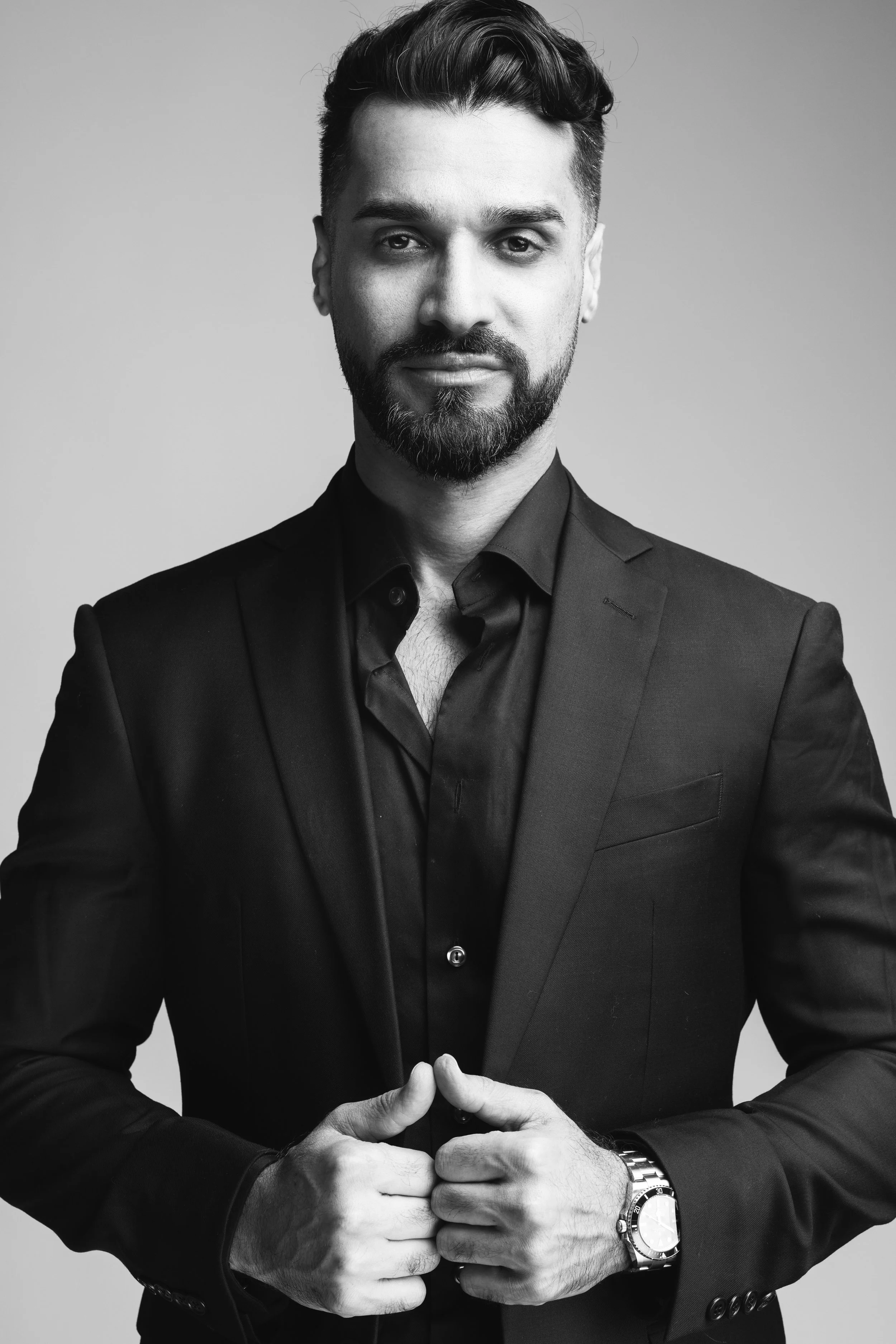 A black and white portrait of a man with dark hair and a beard, wearing a dark suit and shirt, looking confidently at the camera with his hands clasped together.