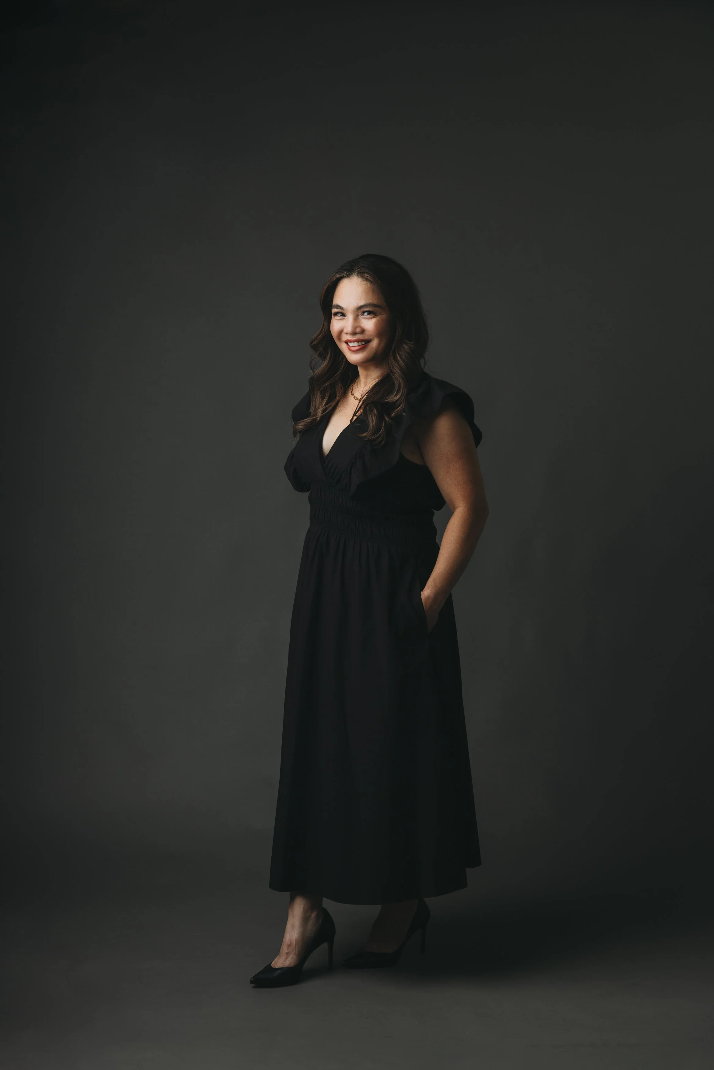 A woman with long wavy brown hair wearing a black dress with ruffled shoulders and high heels, standing against a dark gray background.