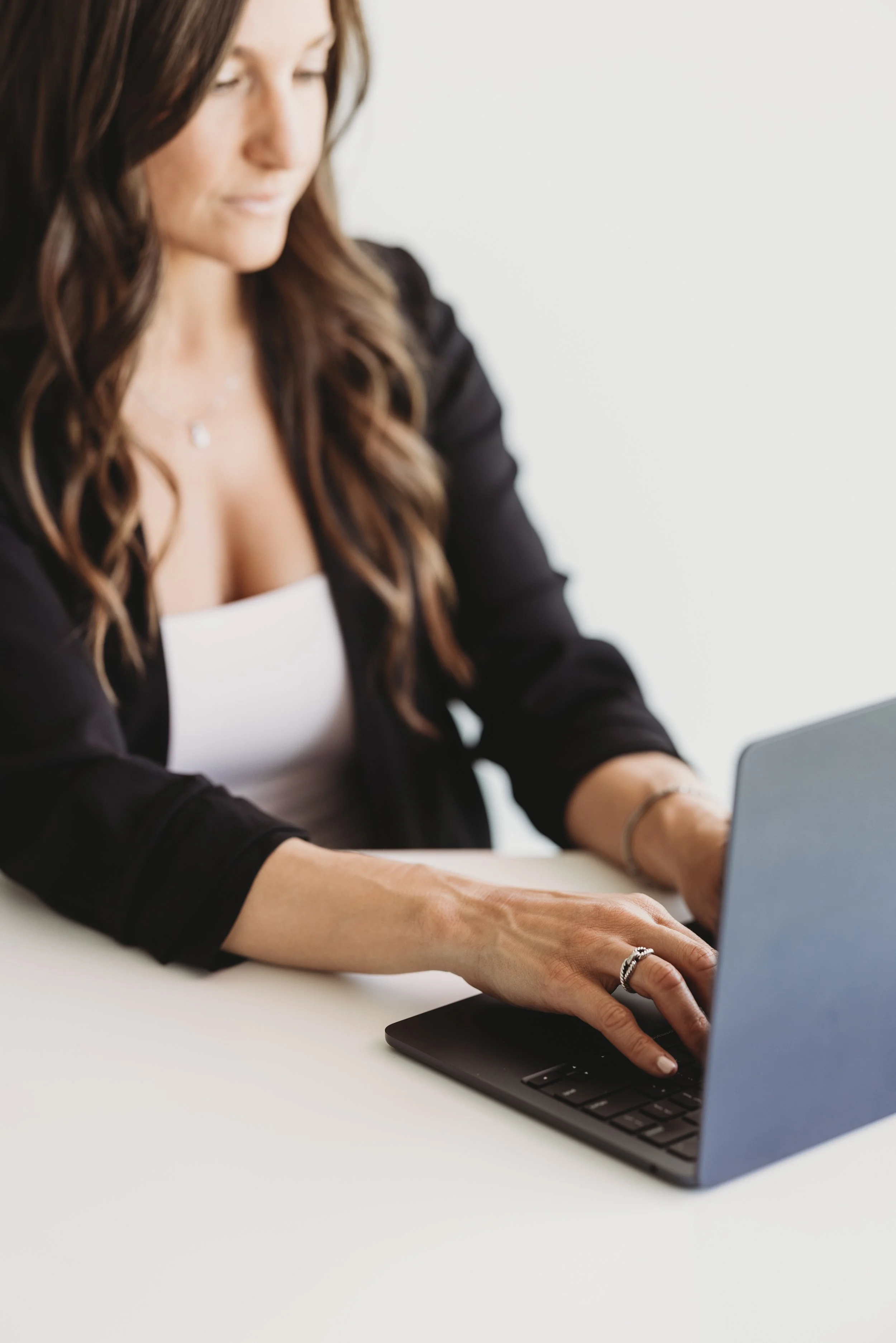 Woman with long wavy hair wearing a black blazer and white top working on a laptop at a white table.