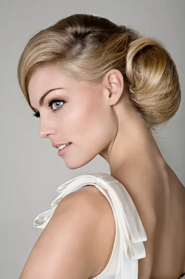 A woman with blonde hair styled in vintage curls, wearing a white dress with ruffled shoulder details, looking to her left with a slight smile.