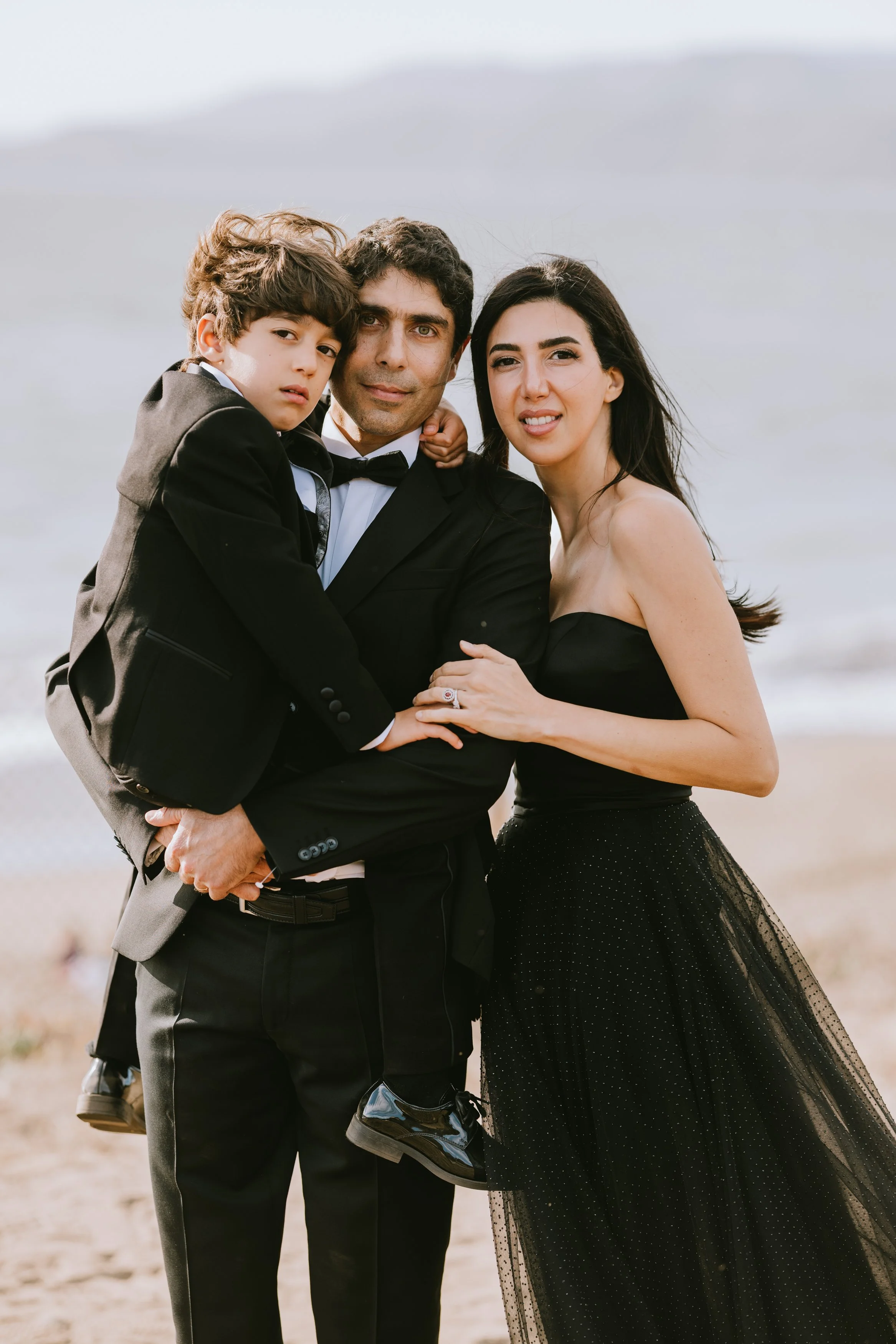 Family of three dressed in formal attire, standing on a beach with sandy ground and a cloudy sky, looking at the camera.