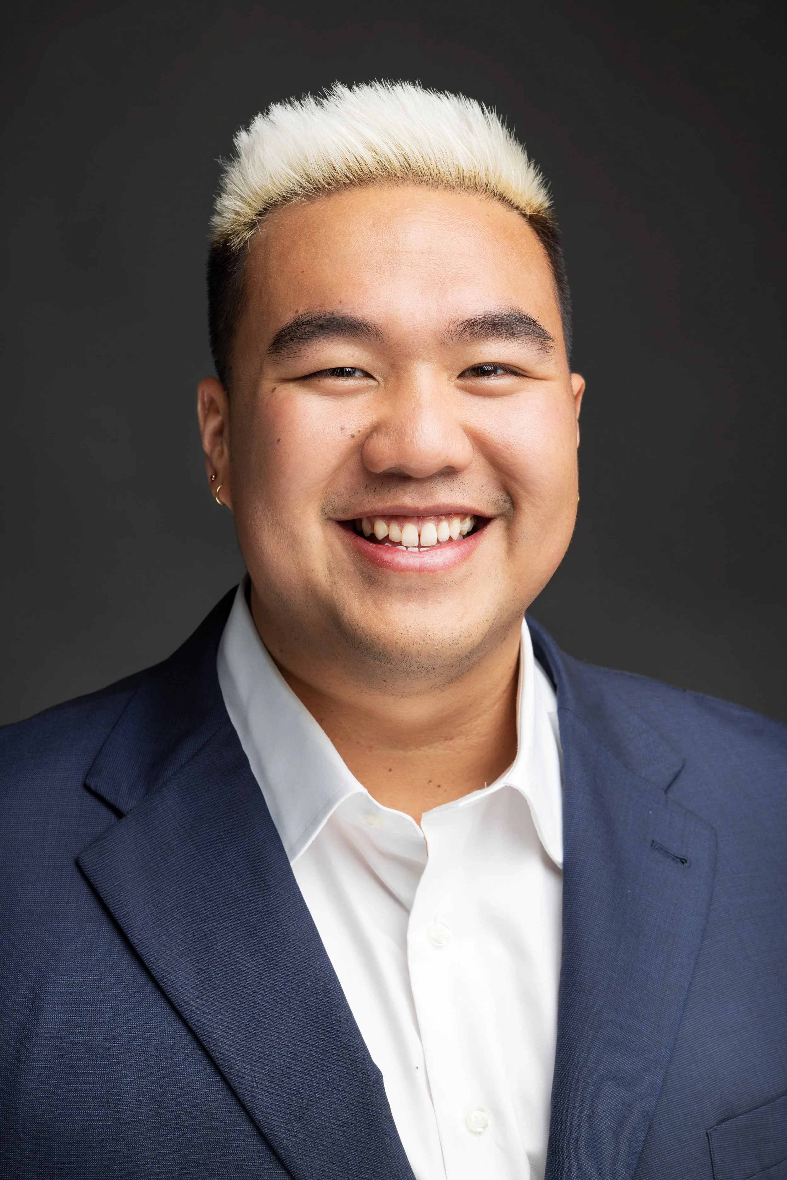 A smiling man with bleached blond hair, wearing a dark blue suit and white shirt, against a dark gradient background.