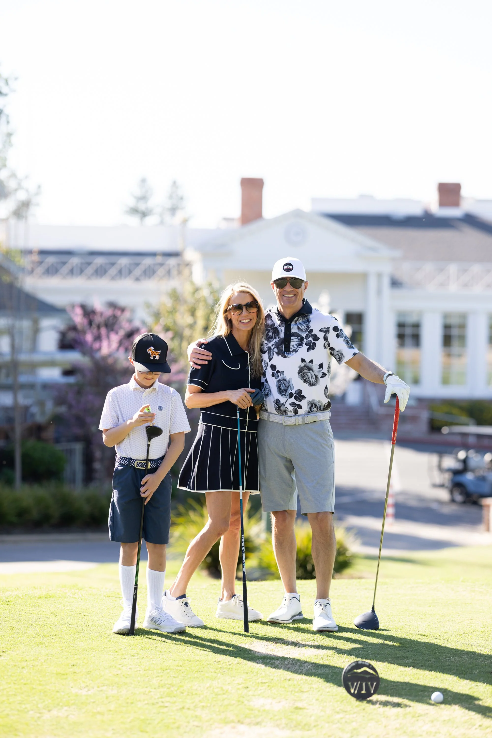 A family of three, two adults and a boy, smiling on a golf course with a large white house in the background. They are dressed in golf attire and holding golf clubs.