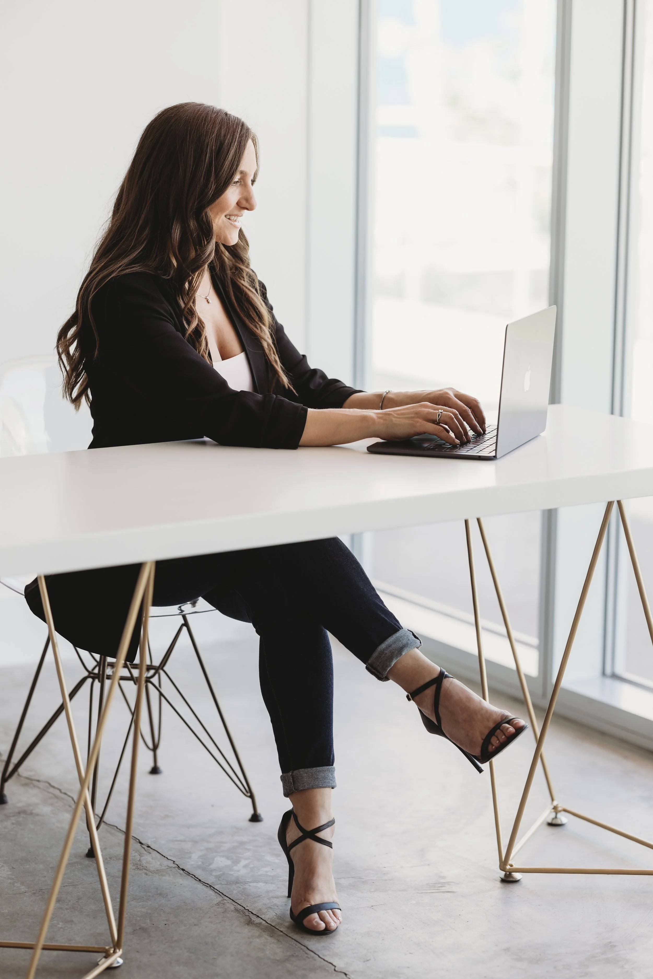 A woman sitting at a white desk using a laptop in a bright office space, with large windows behind her.