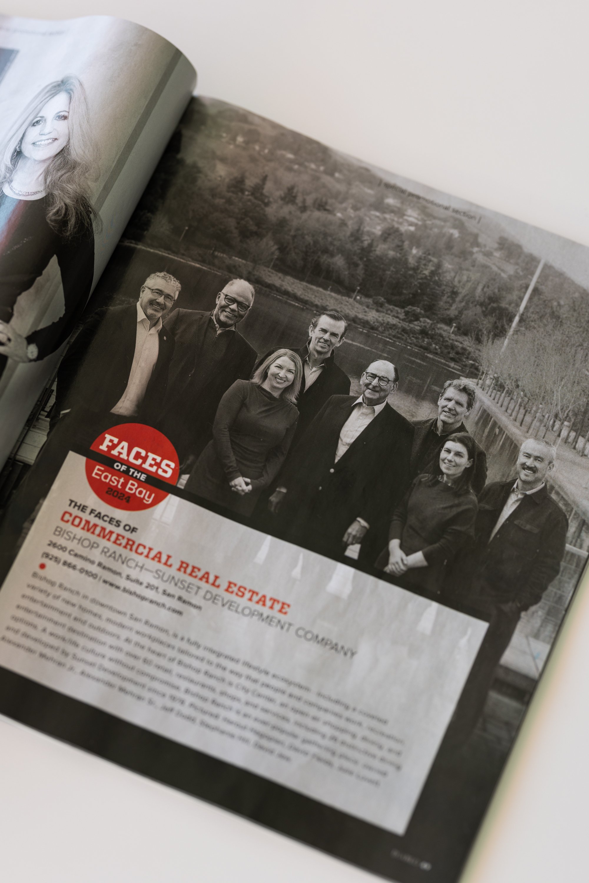 Open magazine featuring a group photo of seven people outdoors with a body of water and trees in the background. The page includes a red and white logo for 'FACES OF THE East Bay' and headlines about real estate and community events.