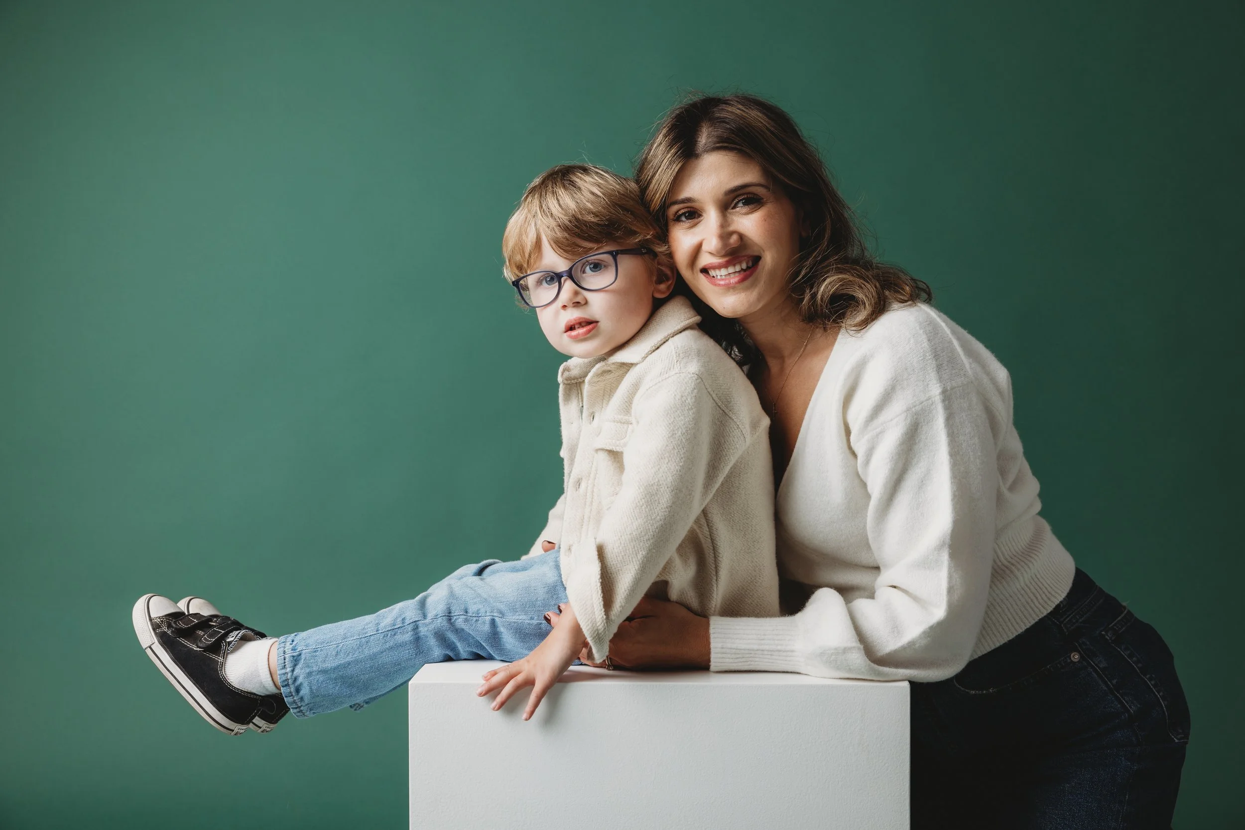 A woman with brown hair smiling while holding a young boy with glasses and red hair, sitting on a white surface against a green background.