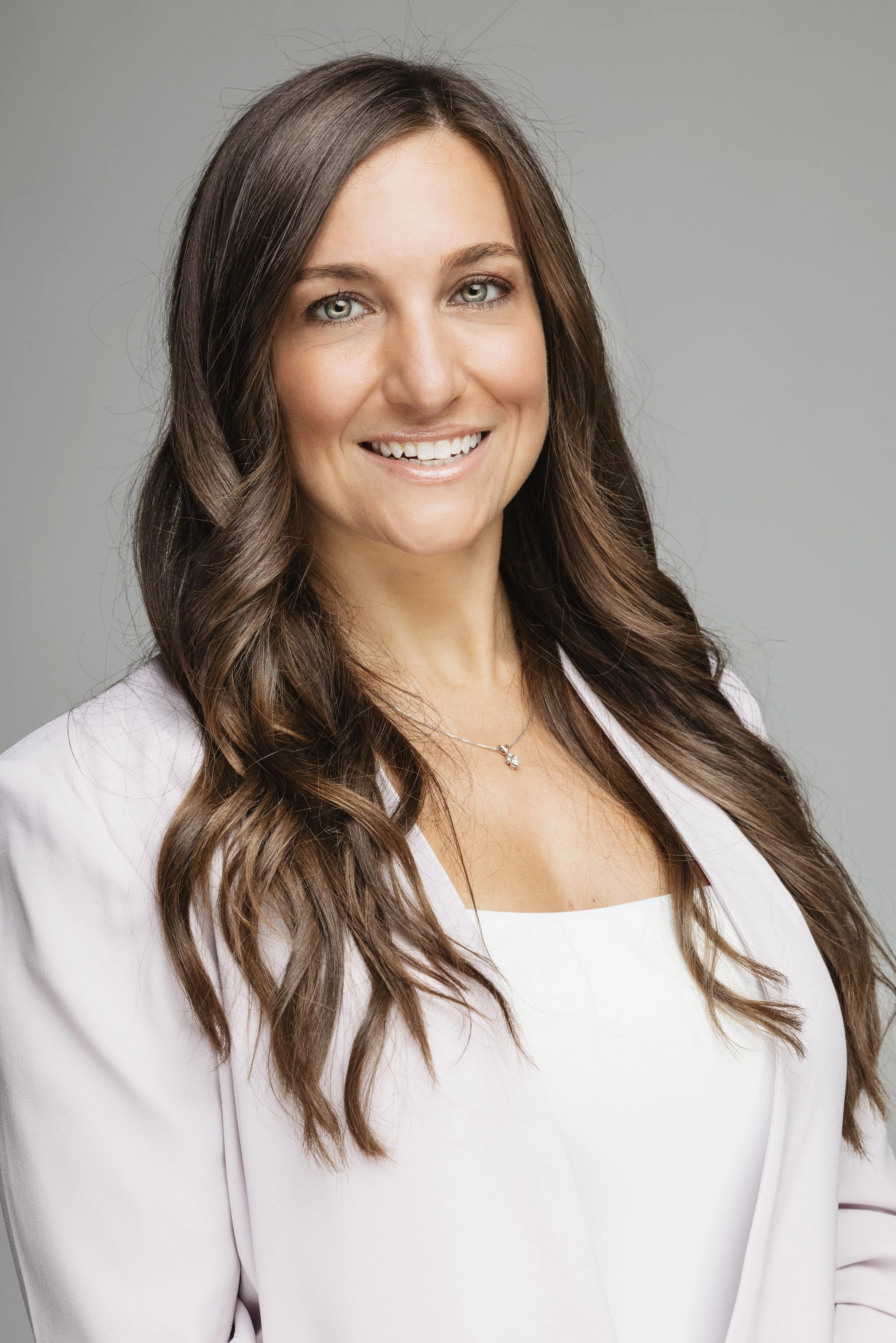 A woman with long, wavy brown hair and green eyes smiling, wearing a white top and a light-colored blazer, with a gray background.