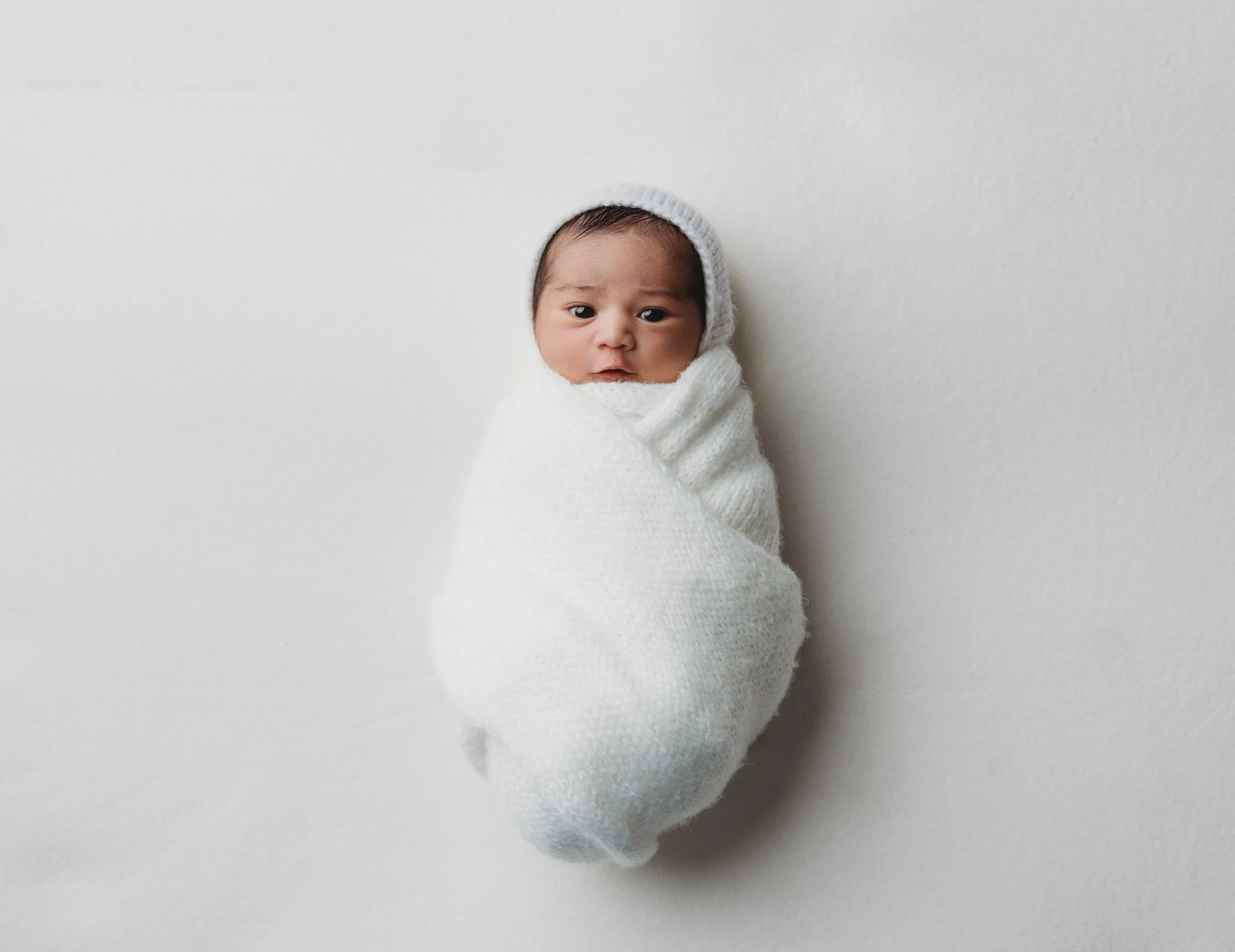 Newborn baby wrapped in white knitted blanket and wearing a white hat, lying on a white surface.