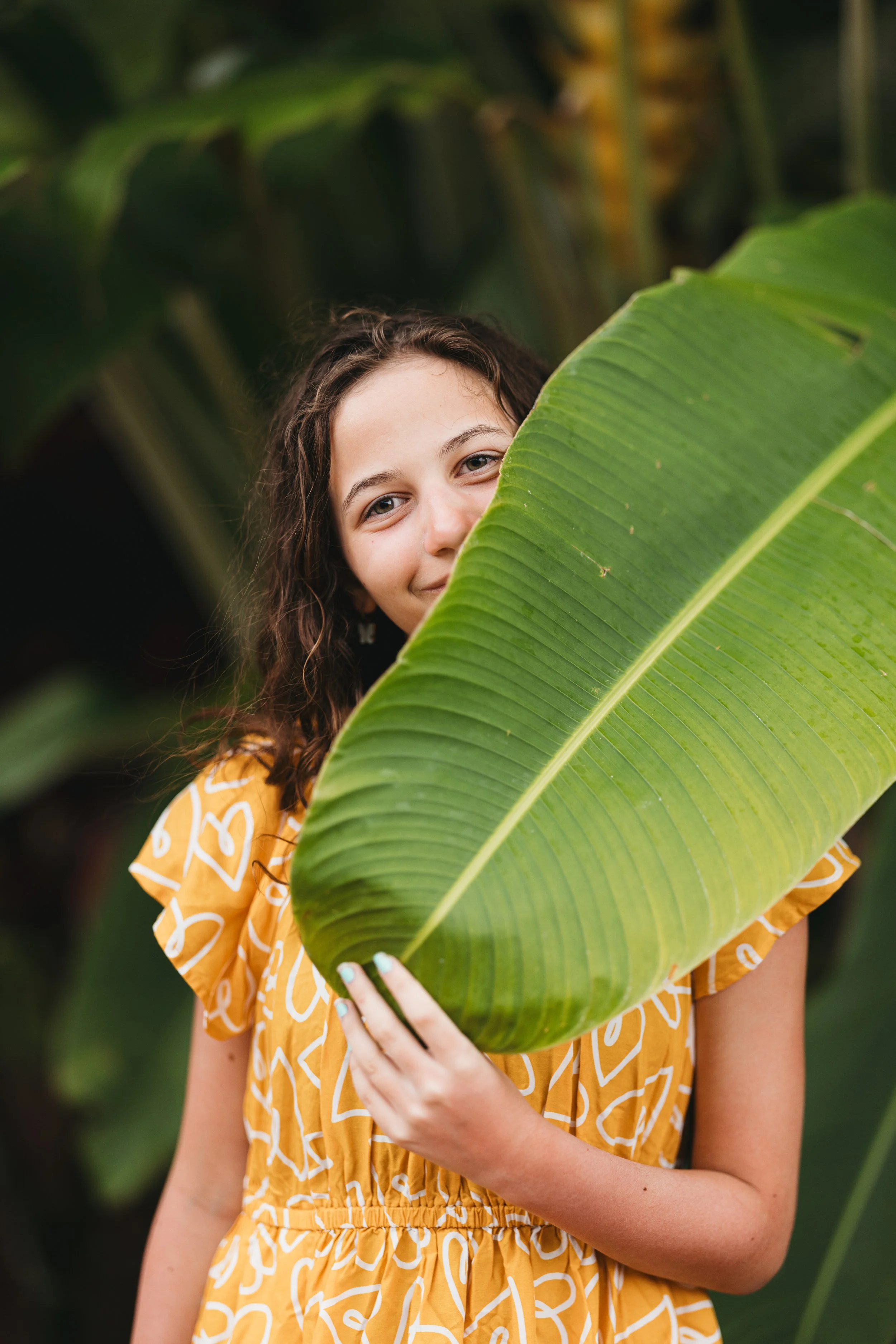 A young girl with curly brown hair and blue eyes peeks from behind a large green banana leaf, wearing a yellow dress with white abstract patterns, in a lush outdoor setting.