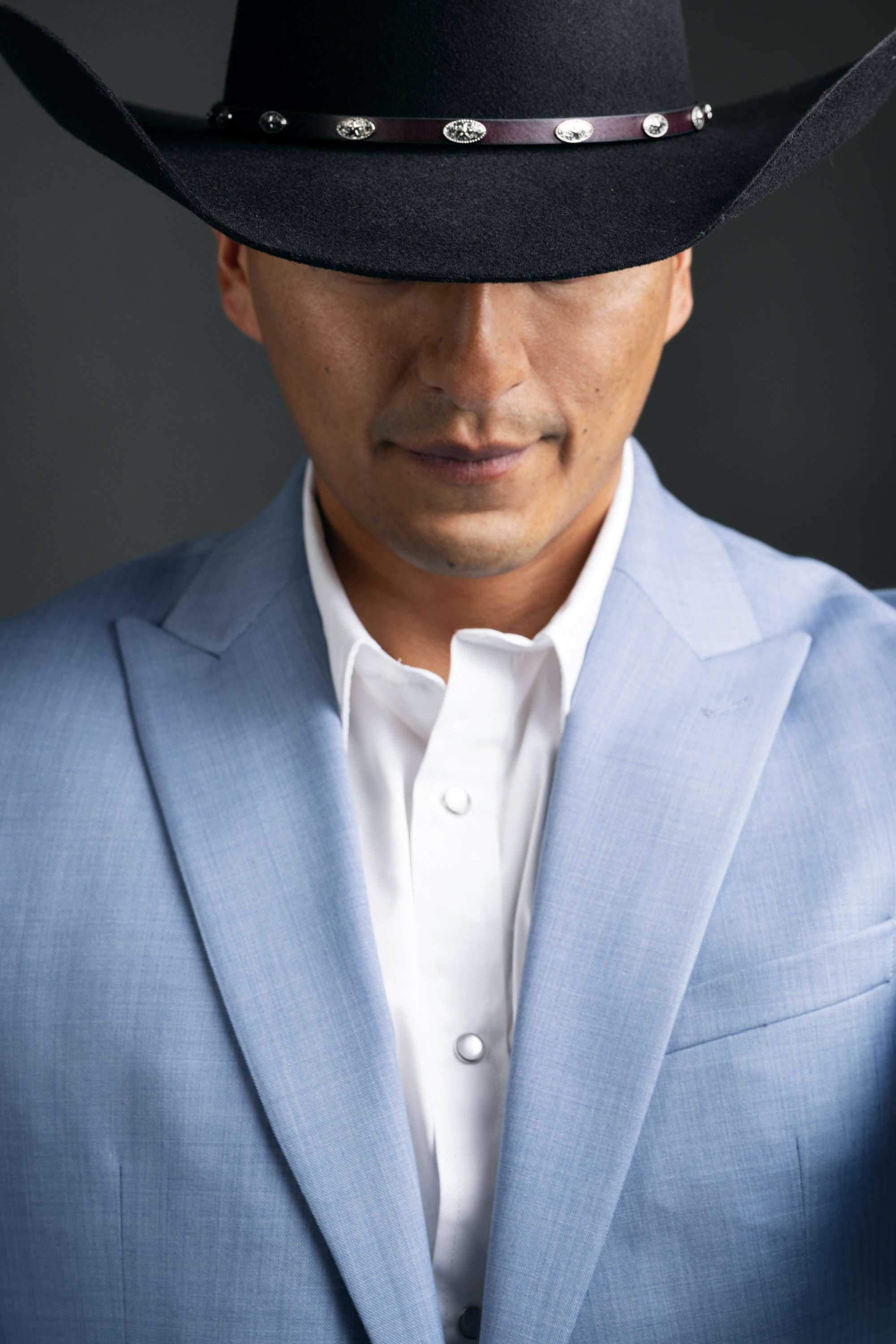 A man wearing a black cowboy hat with decorative silver studs, a light blue blazer, and a white dress shirt, looking downward with a subtle smile.