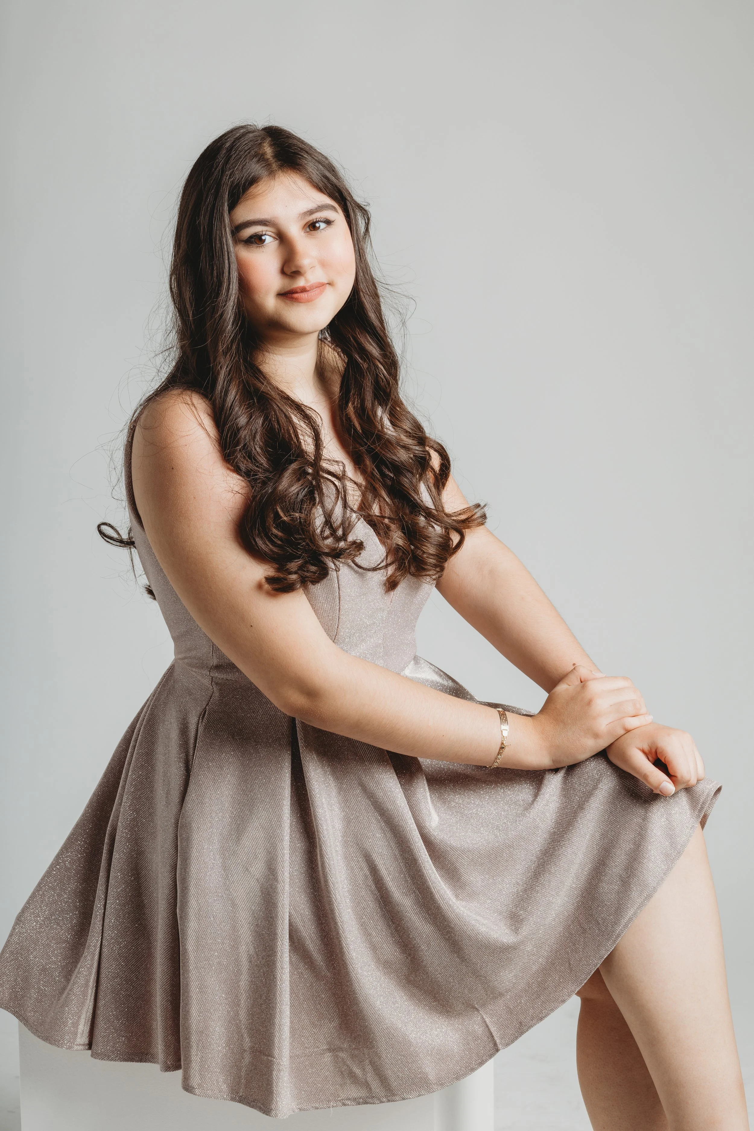 Young woman with long, wavy brown hair sitting on a white stool against a plain white wall, wearing a sleeveless, shimmery beige dress, looking at the camera with a gentle expression.