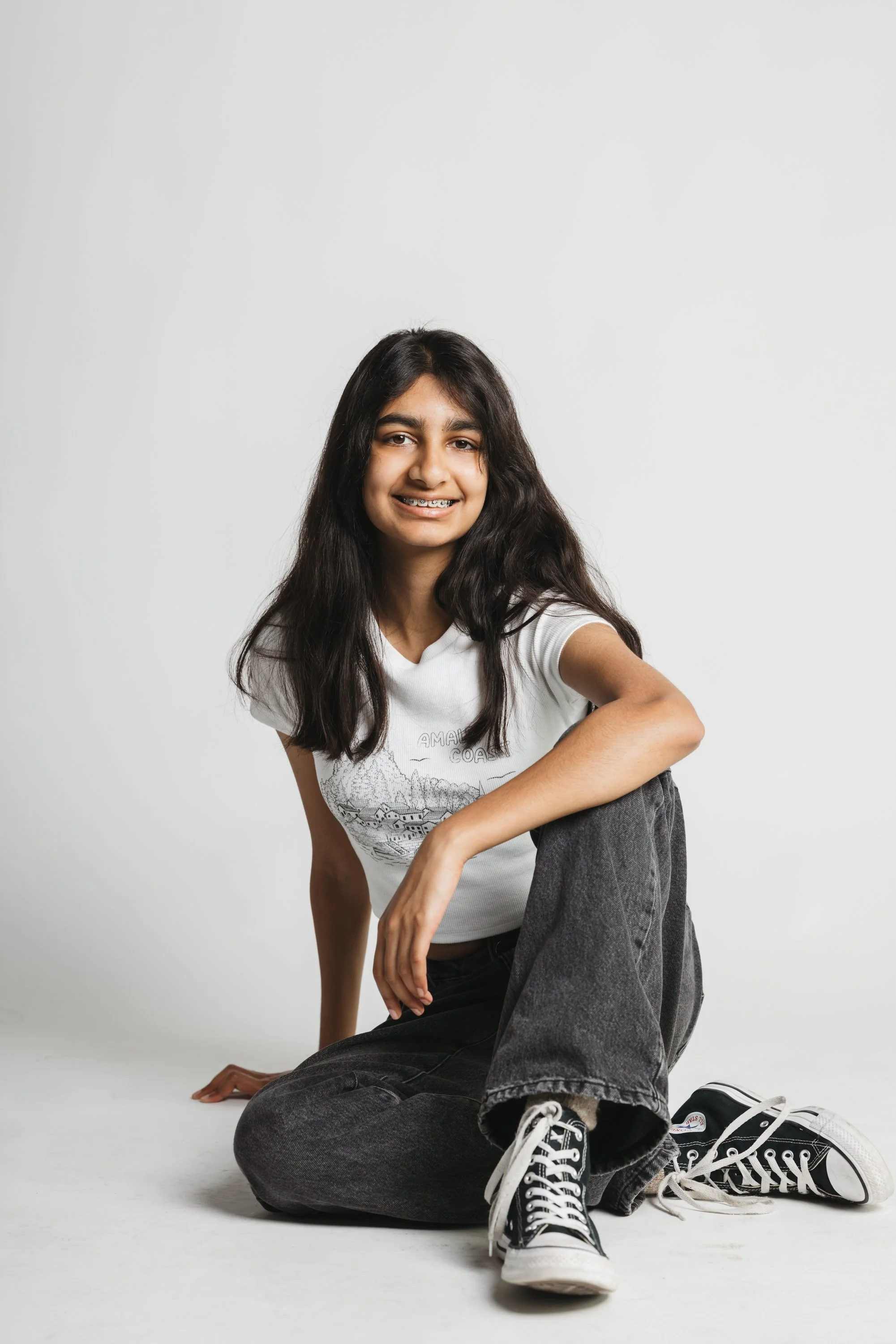 A young girl with long dark hair sitting on the floor, wearing a white t-shirt, dark jeans, and black high-top sneakers, smiling at the camera against a plain white background.