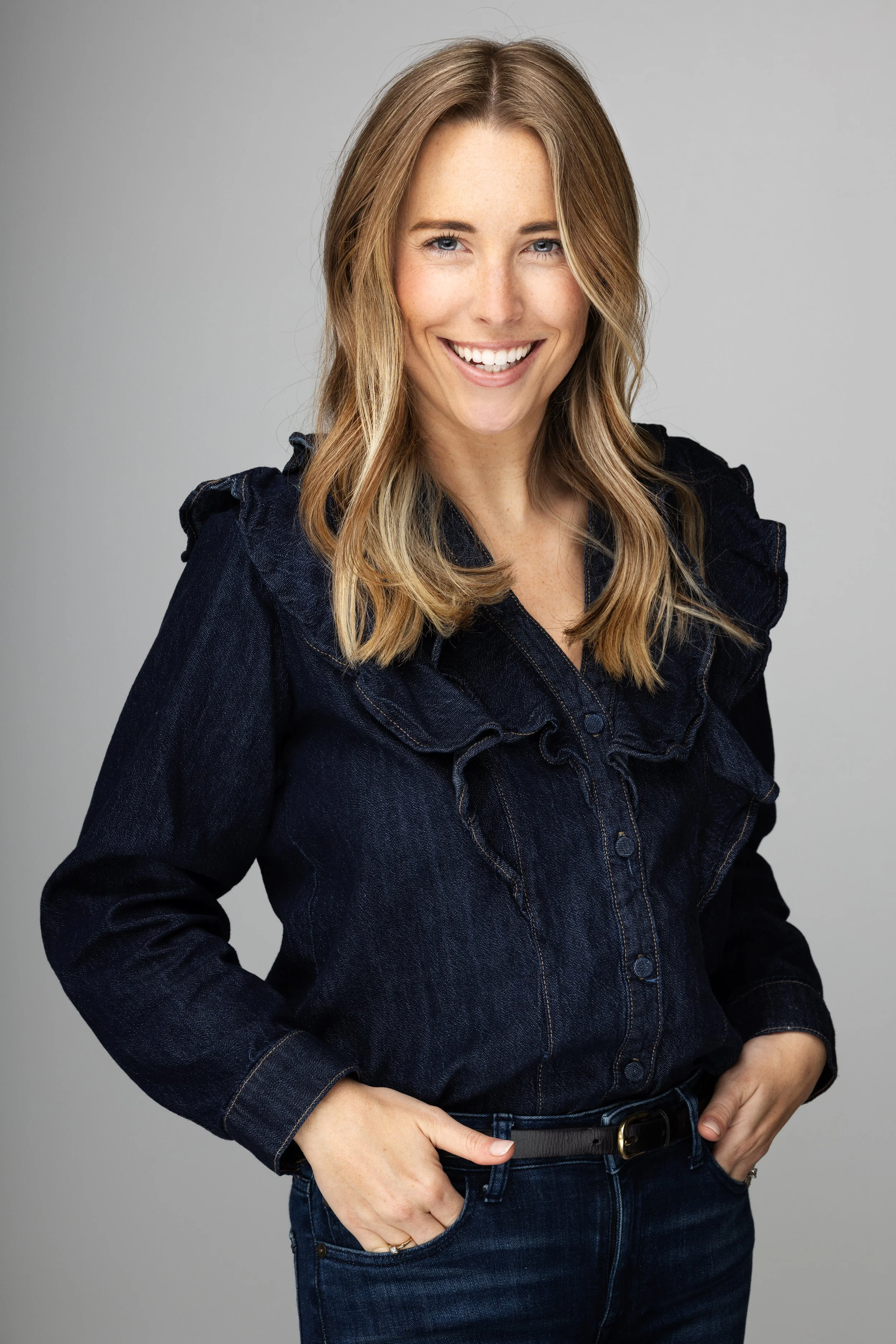 A smiling woman with blonde hair in a black denim jacket against a gray background.