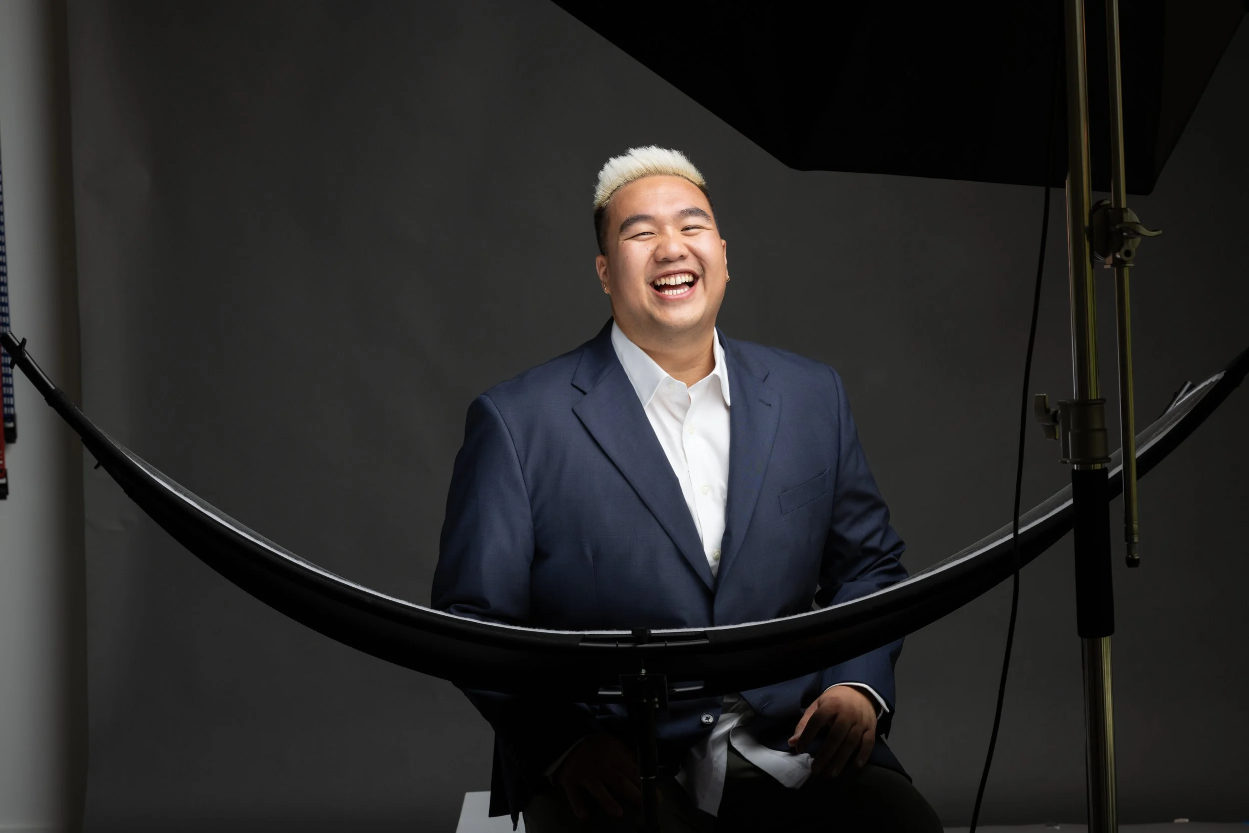 A man with light-colored hair in a navy suit and white shirt smiling and sitting in front of a dark gray backdrop during a photoshoot.